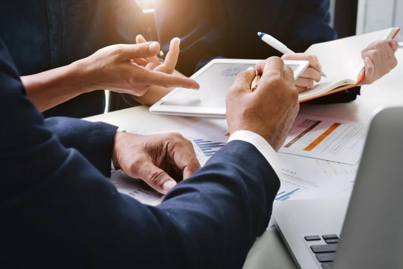 Two professionals discussing documents and data on a table, with a laptop and a tablet, in a business meeting.