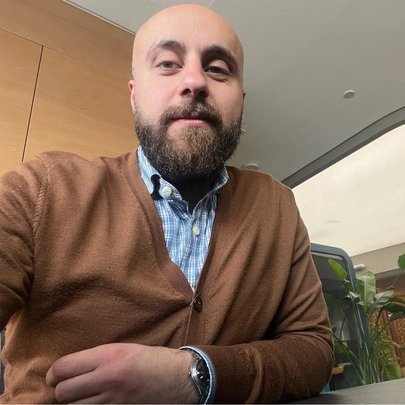 A man with a beard and bald head wearing a brown cardigan and a blue checkered shirt, sitting at a desk with a plant and a computer in a modern office.