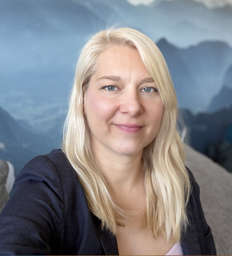 A woman with long blonde hair smiling outdoors with green foliage in the background.