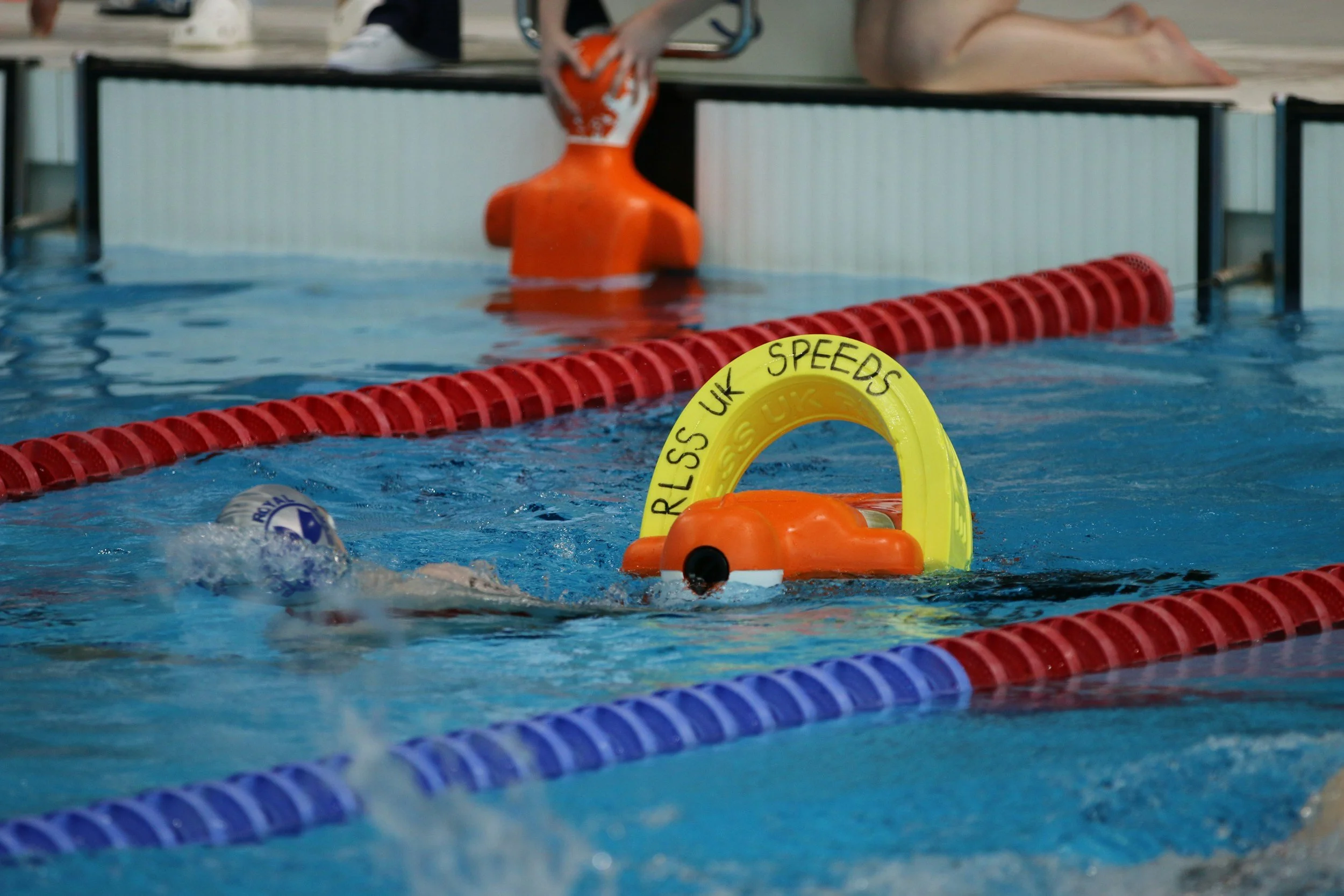 A swimmer in a swimming pool near a yellow and orange lane marker with the words 'RISS UK SPEEDS' written on it. The swimmer is wearing a blue swim cap and goggles and is swimming underwater.