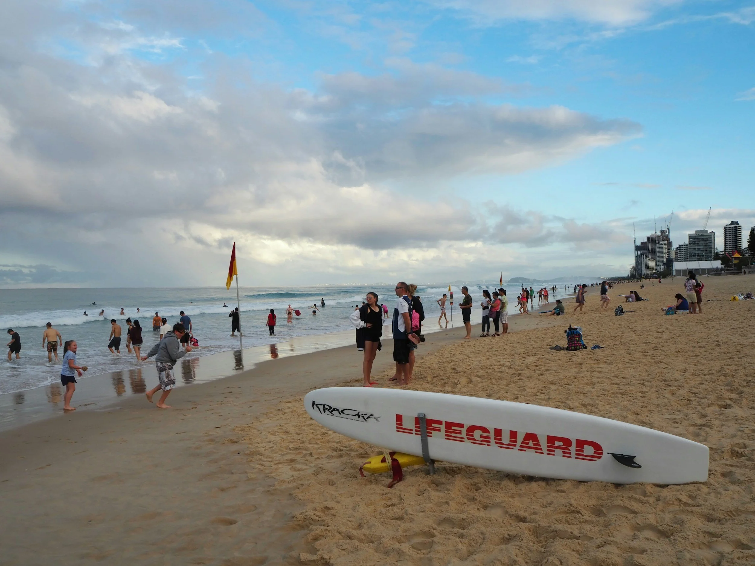 Beach scene with people swimming and relaxing, a surfboard labeled 'LIFEGUARD' on the sand, city buildings in the distance, and cloudy sky above.