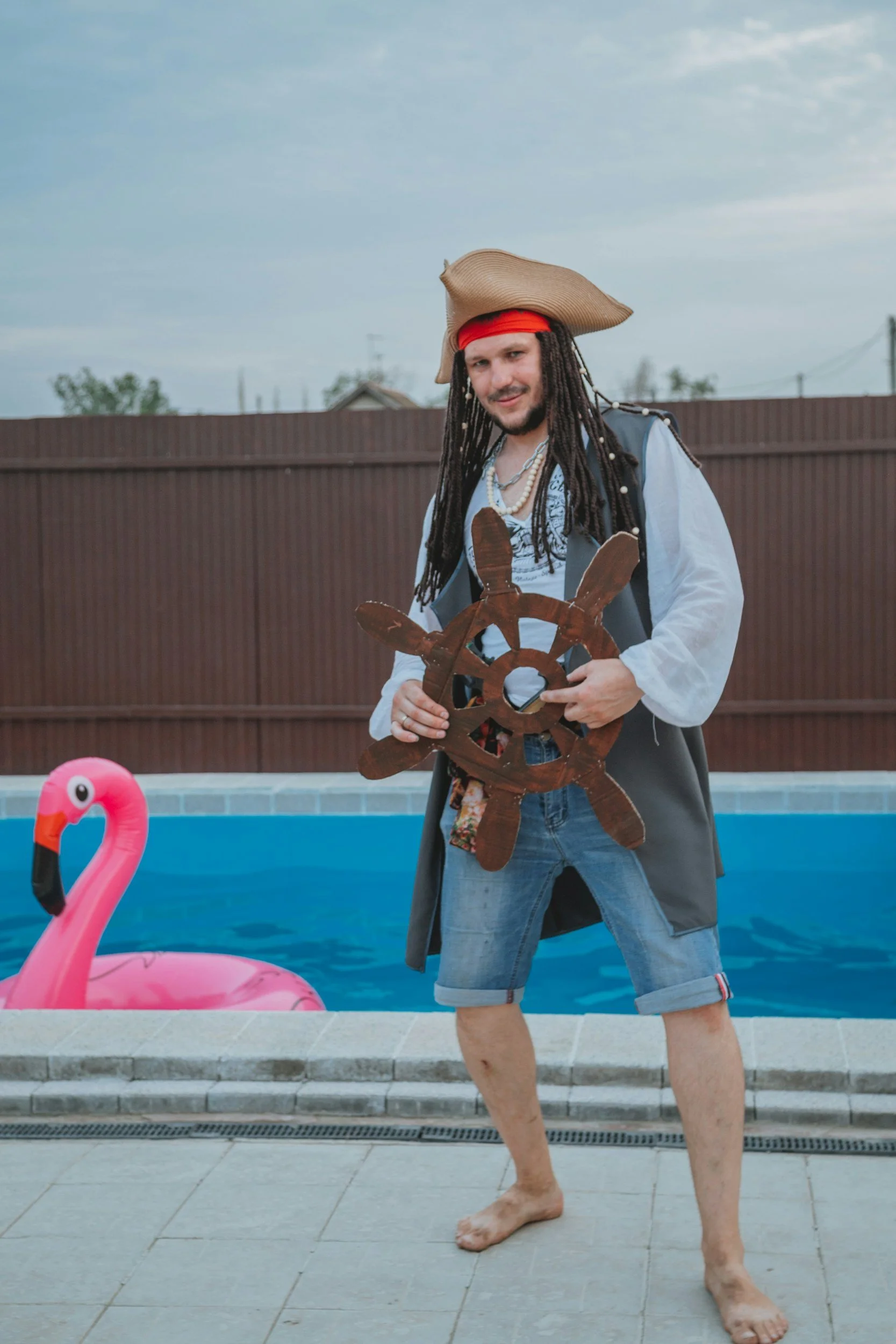 A man dressed as a pirate with a beige hat, red bandana, and dreadlocks, holding a ship's wheel, standing beside a swimming pool with an inflatable pink flamingo float, in an outdoor backyard with a brown fence and cloudy sky.