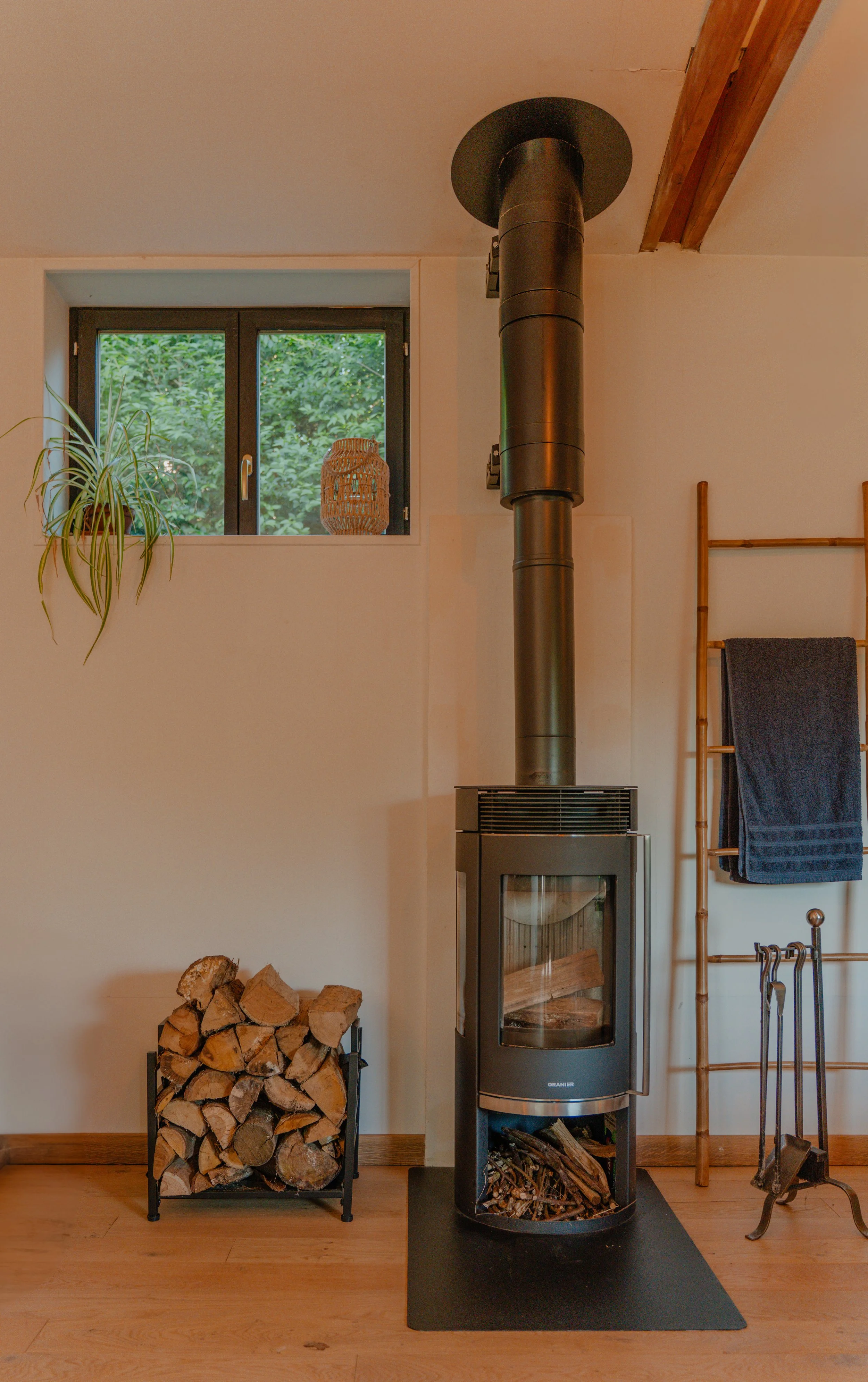 Intérieur d'une pièce avec un poêle en métal, une pile de bois de chauffage, une fenêtre avec vue sur des arbres verts, un panier en osier sur la fenêtre, une étagère en bois avec une serviette ou un tissu sombre, et un porte-rouleau en métal avec outils pour cheminée.