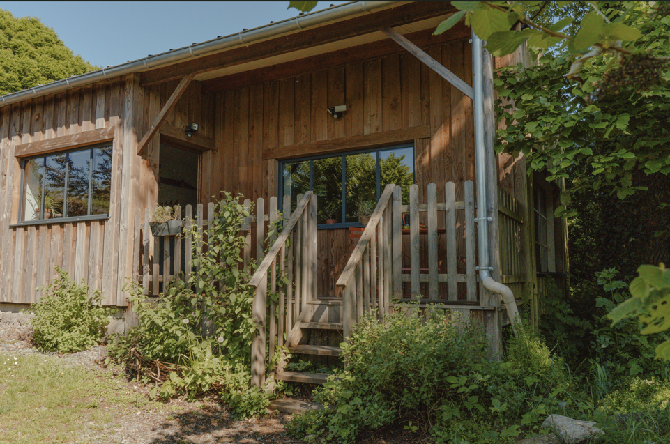 Maison en bois avec petites escaliers menant à une porte, entourée de plantes vertes, avec fenêtres et gouttière visible.