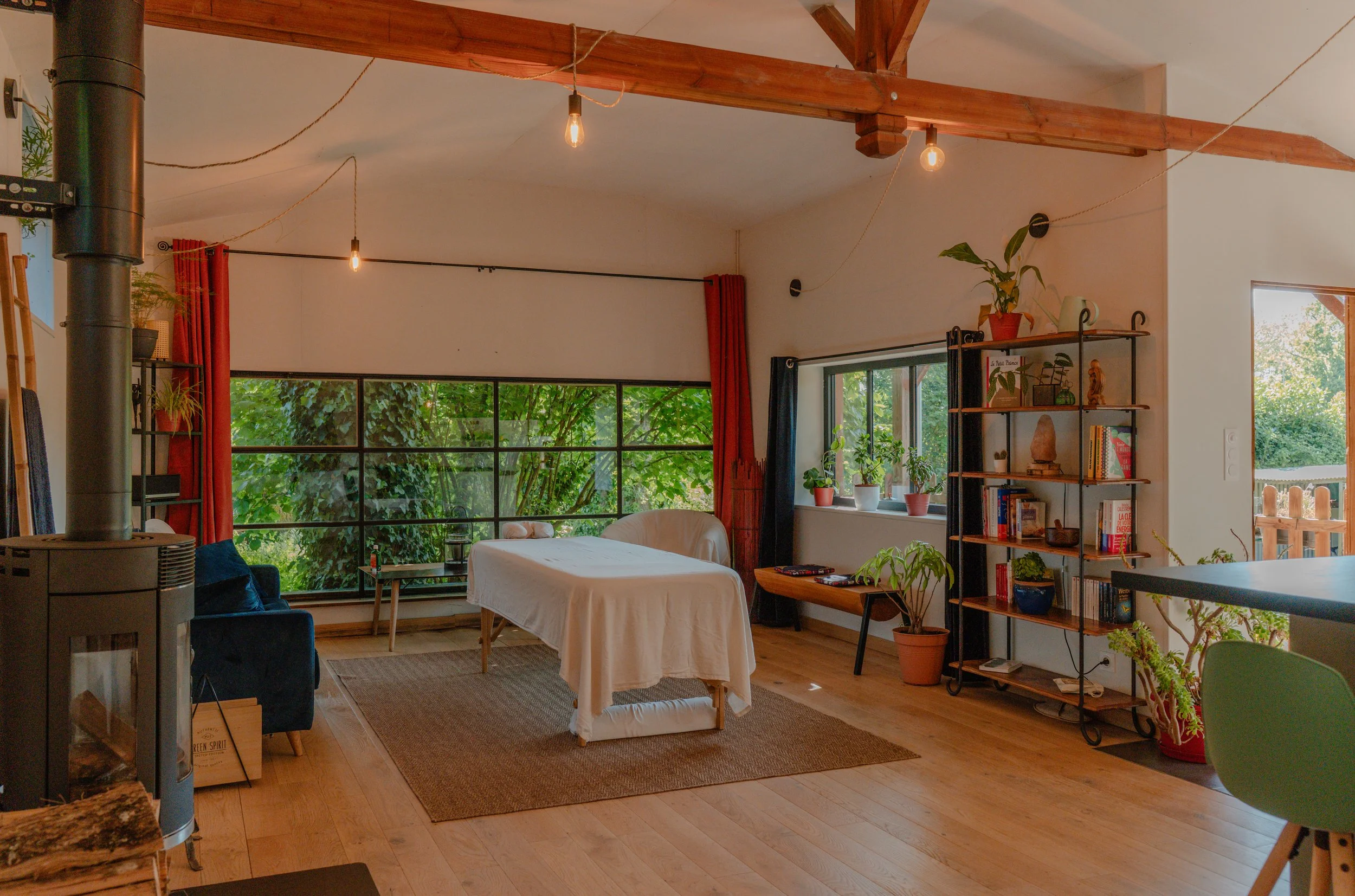 Salon intérieur avec grande fenêtre, espace de vie éclairé, meubles en bois, plantes en pot, étagère avec livres et décorations, rideaux rouges et noirs, plafond avec poutres en bois, lampes à ampoules suspendues.