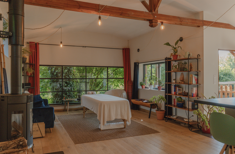 Salon avec grandes fenêtres, table drapée, étagère avec livres et plantes, canapé bleu, cheminée