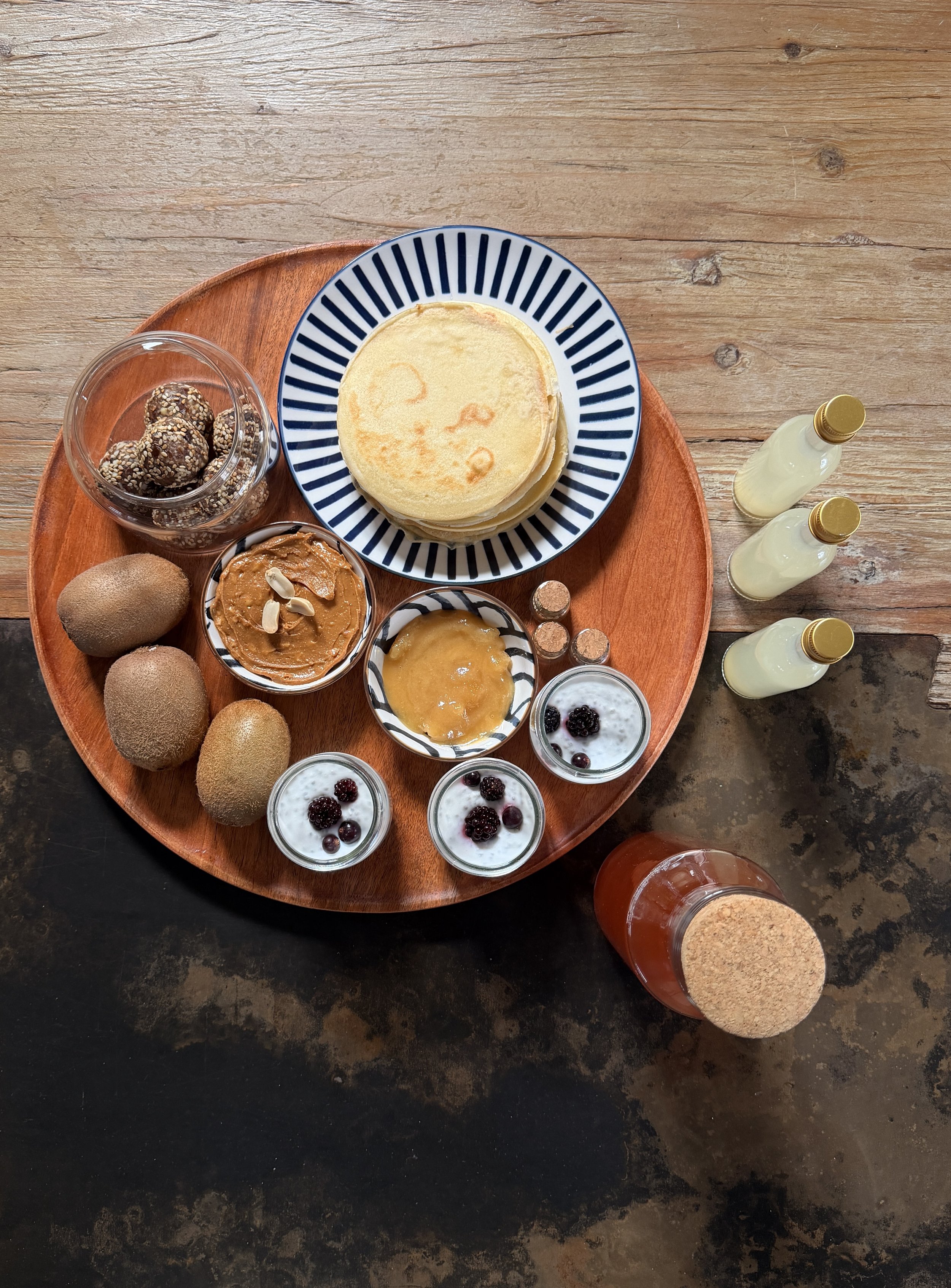 petit déjeuner fait maison, Un plateau en bois avec différentes collations et boissons. Il y a des crêpes empilées, des kiwis, du beurre de cacahuète, du yaourt avec des mûres, des petites bouteilles de lait ou sirop, et d'autres desserts et snacks.