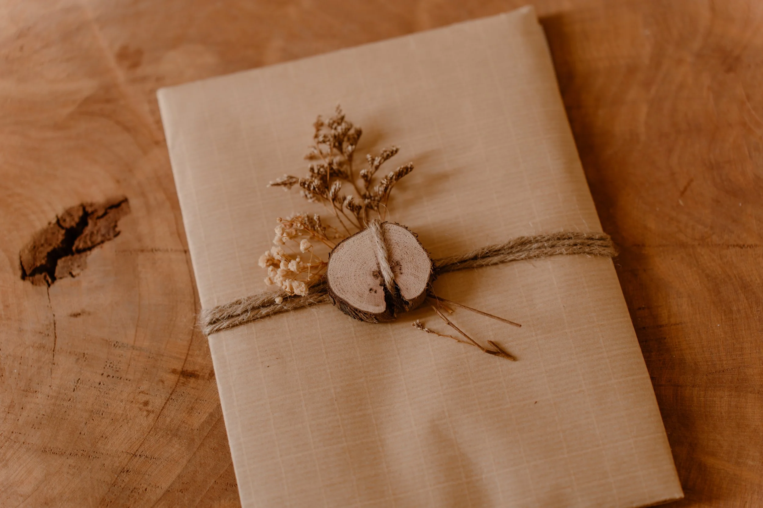 Carnet d'aventure enveloppé dans du papier beige, décoré d'une tranche de bois, de petites plantes séchées, et attaché avec une ficelle pour un envoi naturel.