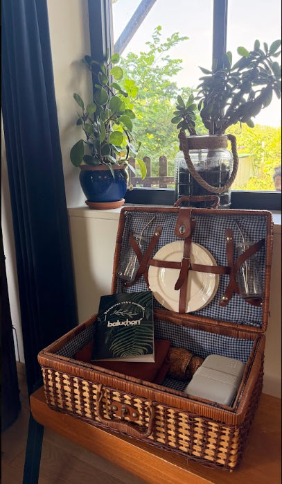 panier pique nique en osier ouverte contenant un livre, un petit coffret, et des verres à vin, placée à côté d'une fenêtre avec des plantes vertes en pot sur le rebord.