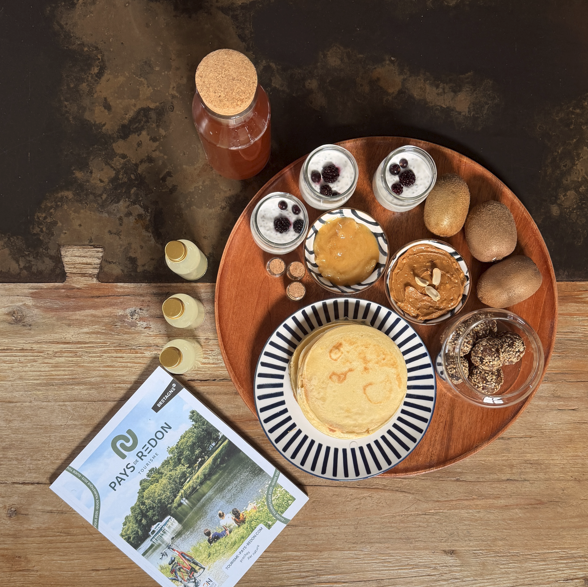 Petit déjeuner avec pancakes, kiwi, confiture, petits pots, sirop, carte touristique de la région Pays de Redon, et autres accompagnements sur un plateau en bois.