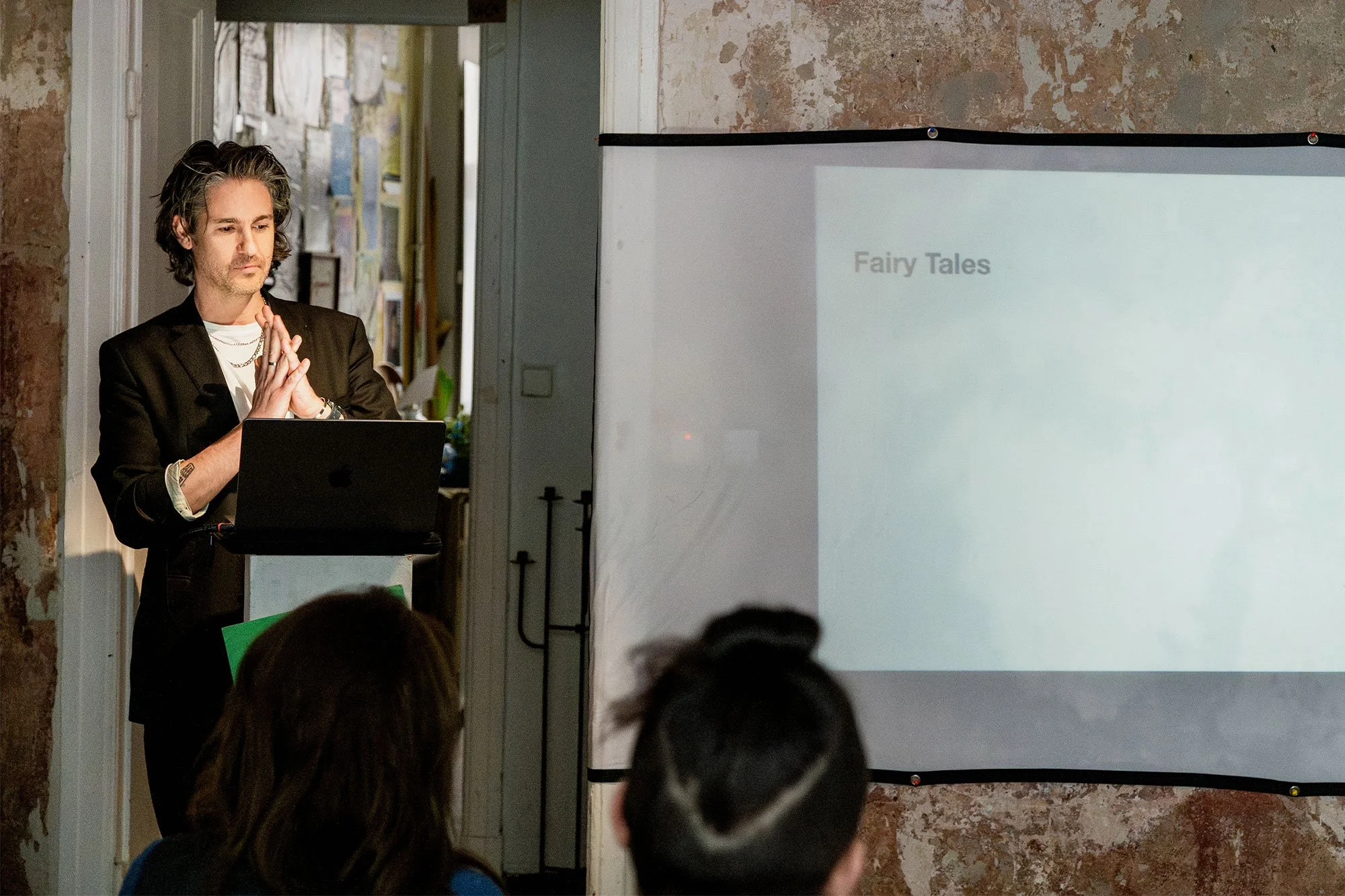 Ein Mann hält eine Präsentation mit einem Laptop vor einer Leinwand, auf der 'Fairy Tales' steht, vor einer Gruppe von Zuhörern in einem Raum mit rohen Wänden.