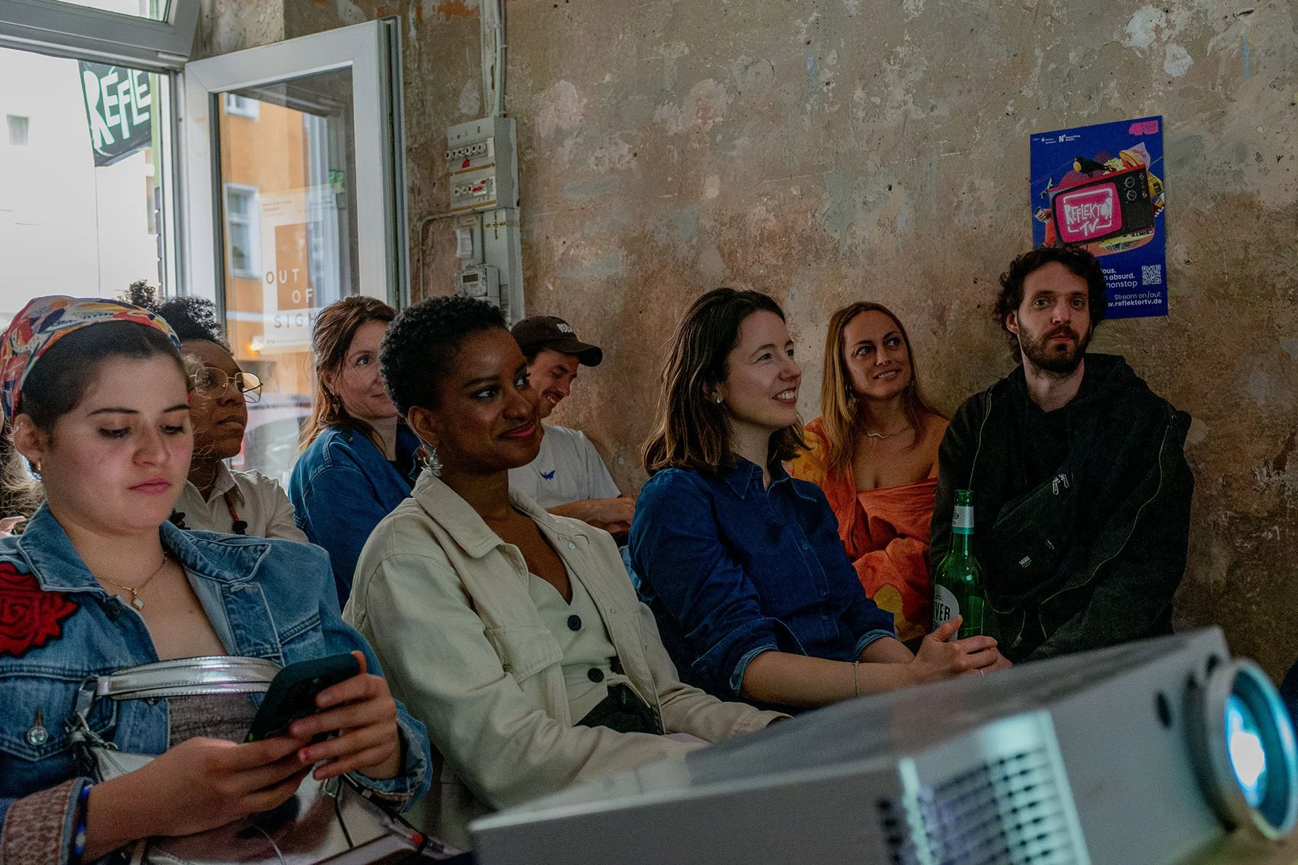Eine Gruppe von acht jungen Erwachsenen sitzt in einem Raum mit rauer Wand und einem Fenster. Sie scheinen einem Vortrag zuzuhören, einige lachen, andere schauen interessiert. Eines der Mädchen ist auf ihr Smartphone konzentriert, während ein Mann eine Flasche Bier hält.