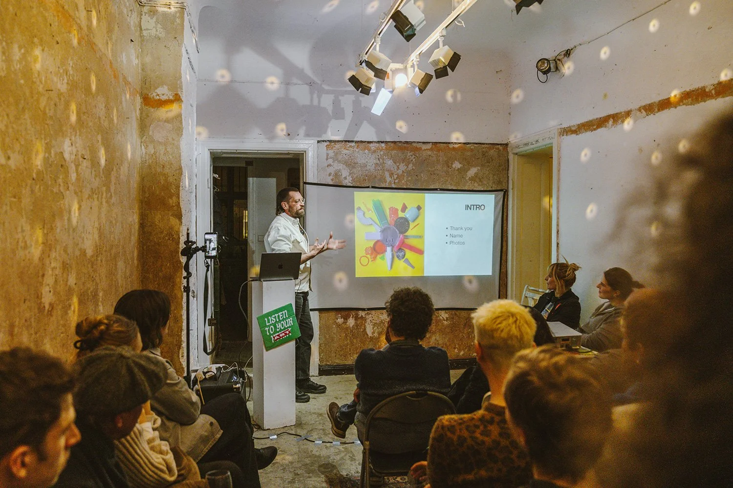 Ein Mann hält eine Präsentation vor einer Gruppe von Menschen in einem Raum mit abblätternden Wandflächen und Professional-Lichter. Es gibt eine Leinwand mit einer Folie, die die Wörter 'Intro', 'Thank you', 'Name' und 'Photos' zeigt.