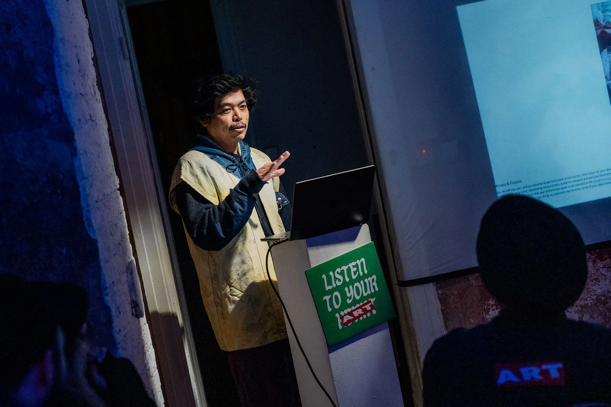 Eine Person steht vor einem Bildschirm, hält eine Hand zum Sprechen und trägt eine Jacke. Im Vordergrund sind Silhouetten von Zuhörern zu sehen, einer trägt ein T-Shirt mit dem Text 'ART'.