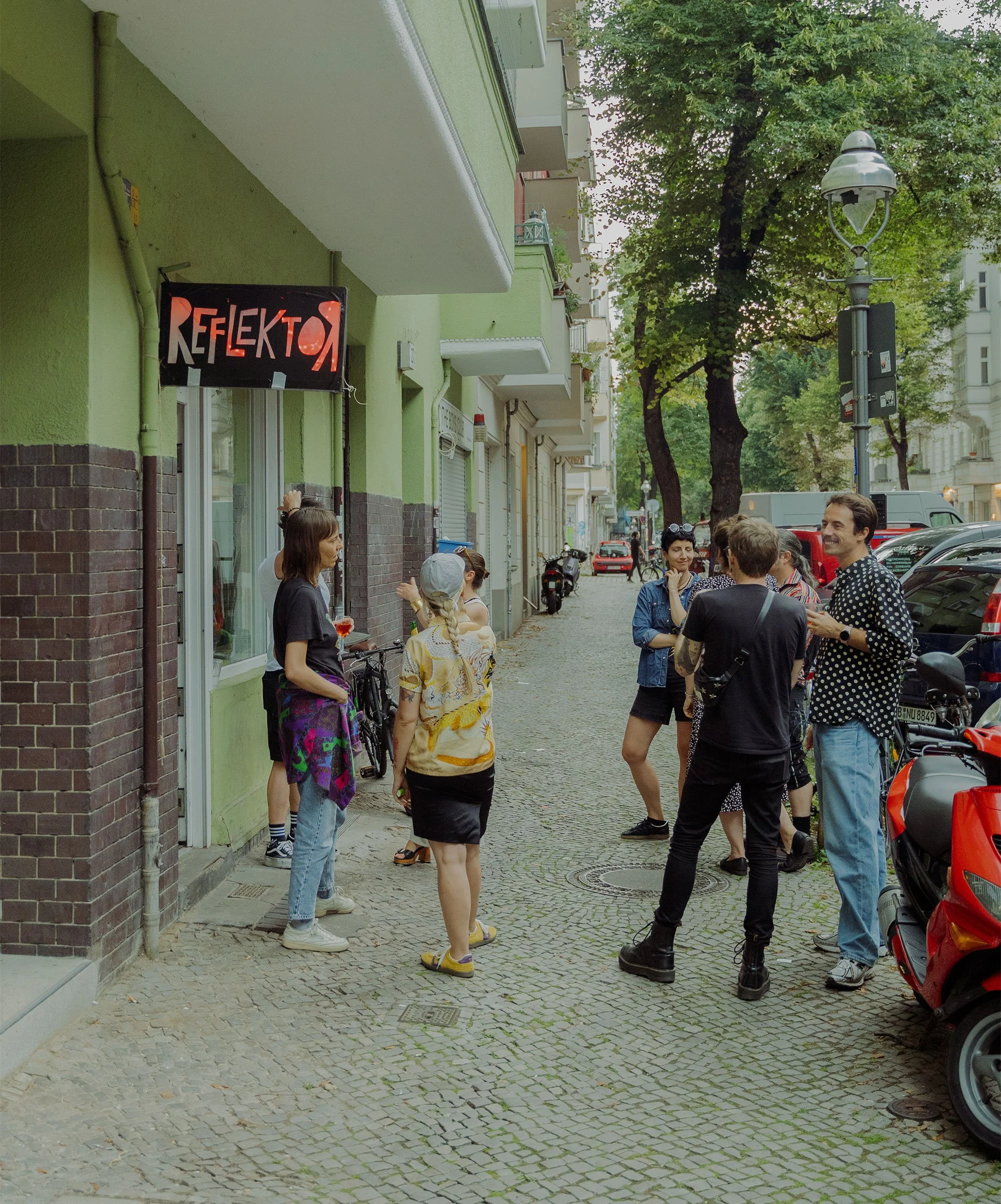 Eine Gruppe von Menschen steht in einer Straße vor einem Laden namens Reflexo, unter einem Baum, im Gespräch, bei Sonnenuntergang.