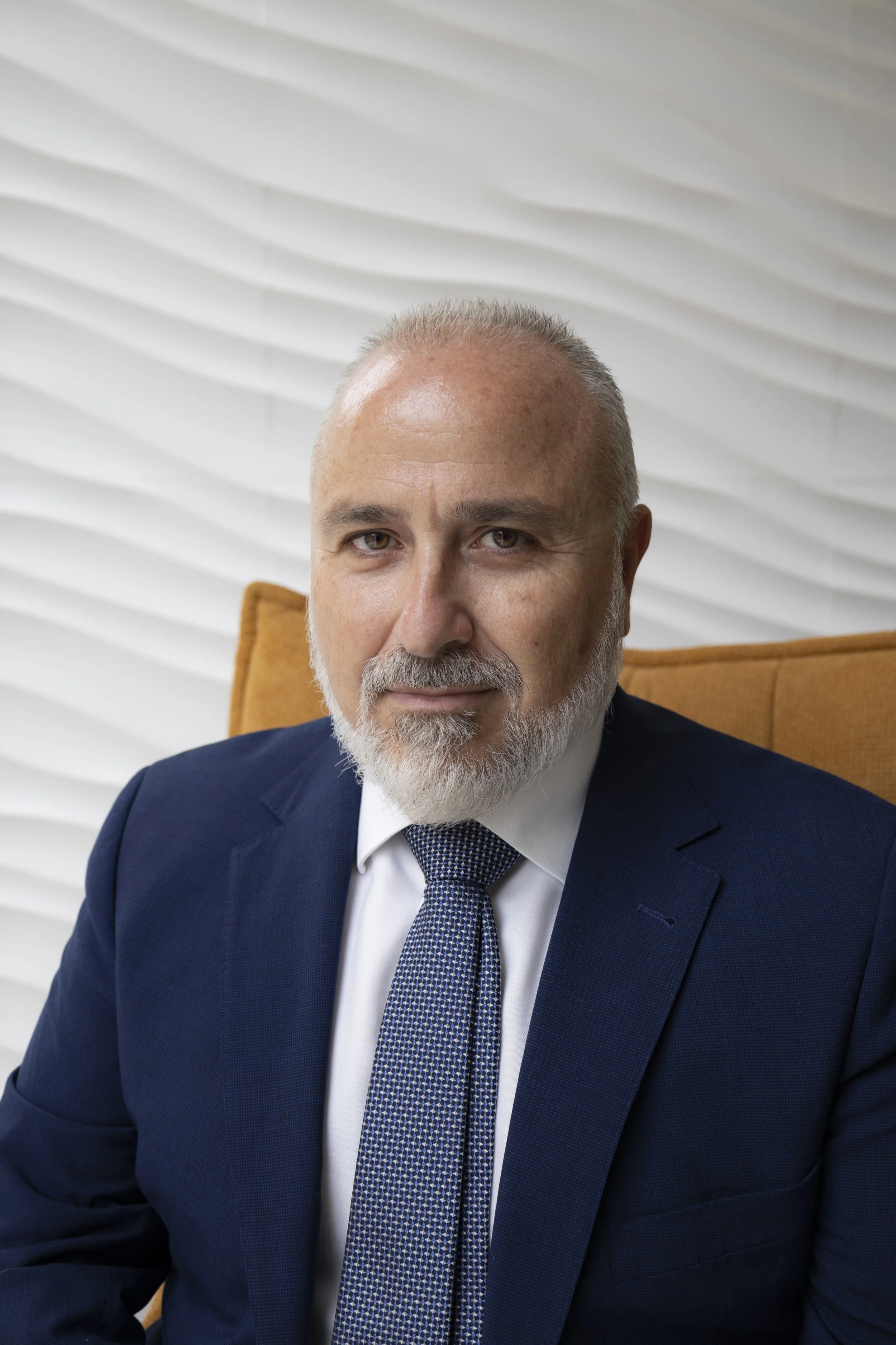 Retrato de un hombre de cabello canoso y barba, vestido con traje azul y corbata, sentado en una silla anaranjada, con fondo de ventanas con persianas blancas.