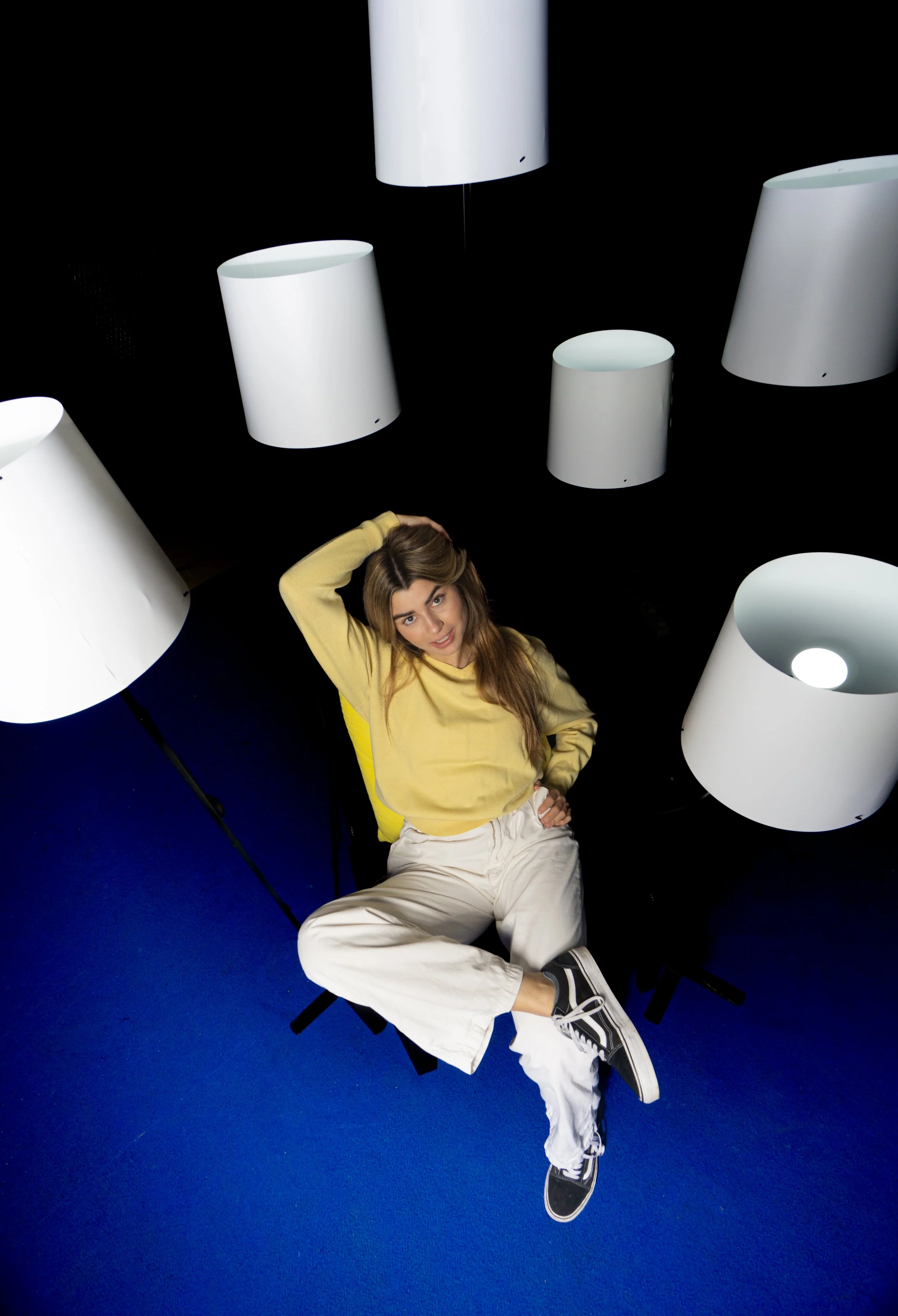 Mujer joven sentada en una silla negra, rodeada de lámparas blancas en un fondo oscuro, vestida con camiseta amarilla y pantalones blancos, con zapatos deportivos negros y blancos.