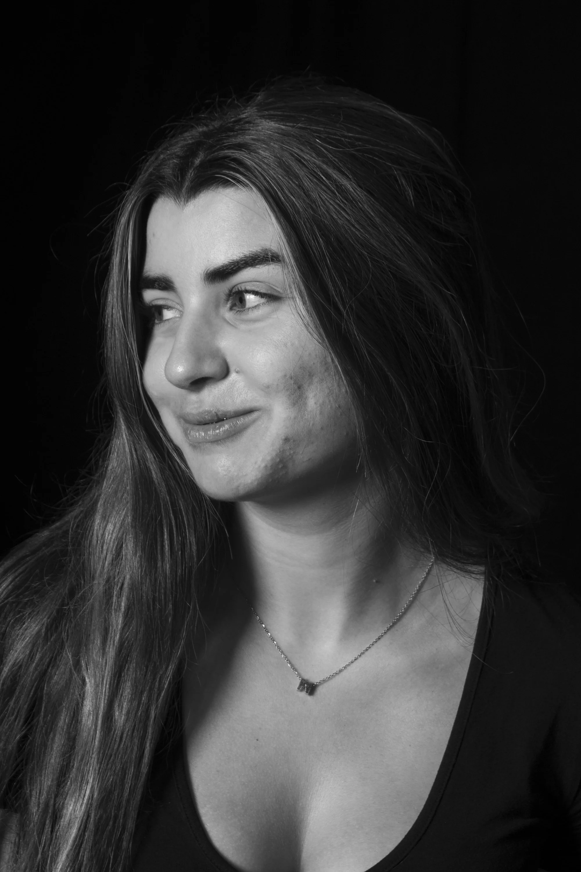 Retrato en blanco y negro de una joven sonriendo, con cabello largo y suelto, usando collar y vestimenta oscura, fondo negro.