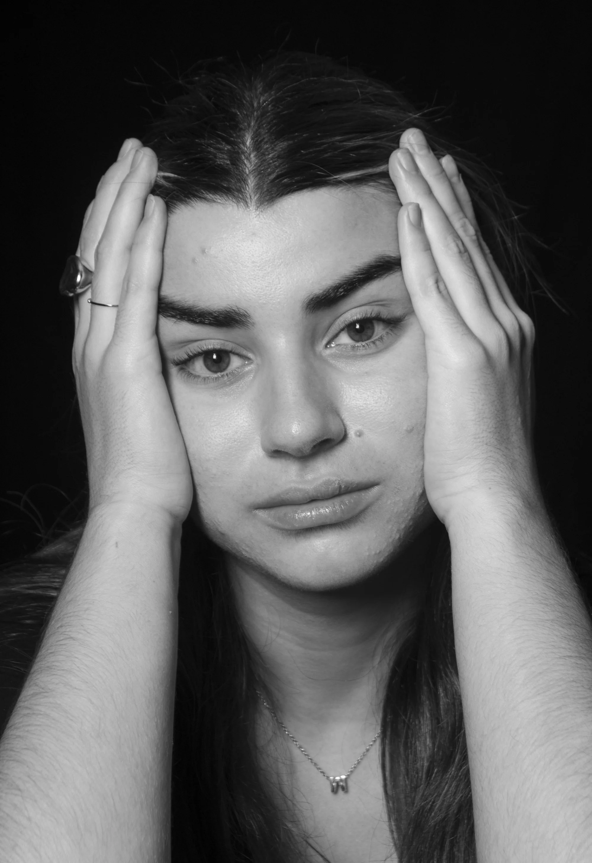 Retrato en blanco y negro de una mujer con expresión pensativa, tocándose la cabeza con ambas manos, cabello largo y liso, usando collar con la letra 'M' y un anillo en la mano izquierda.
