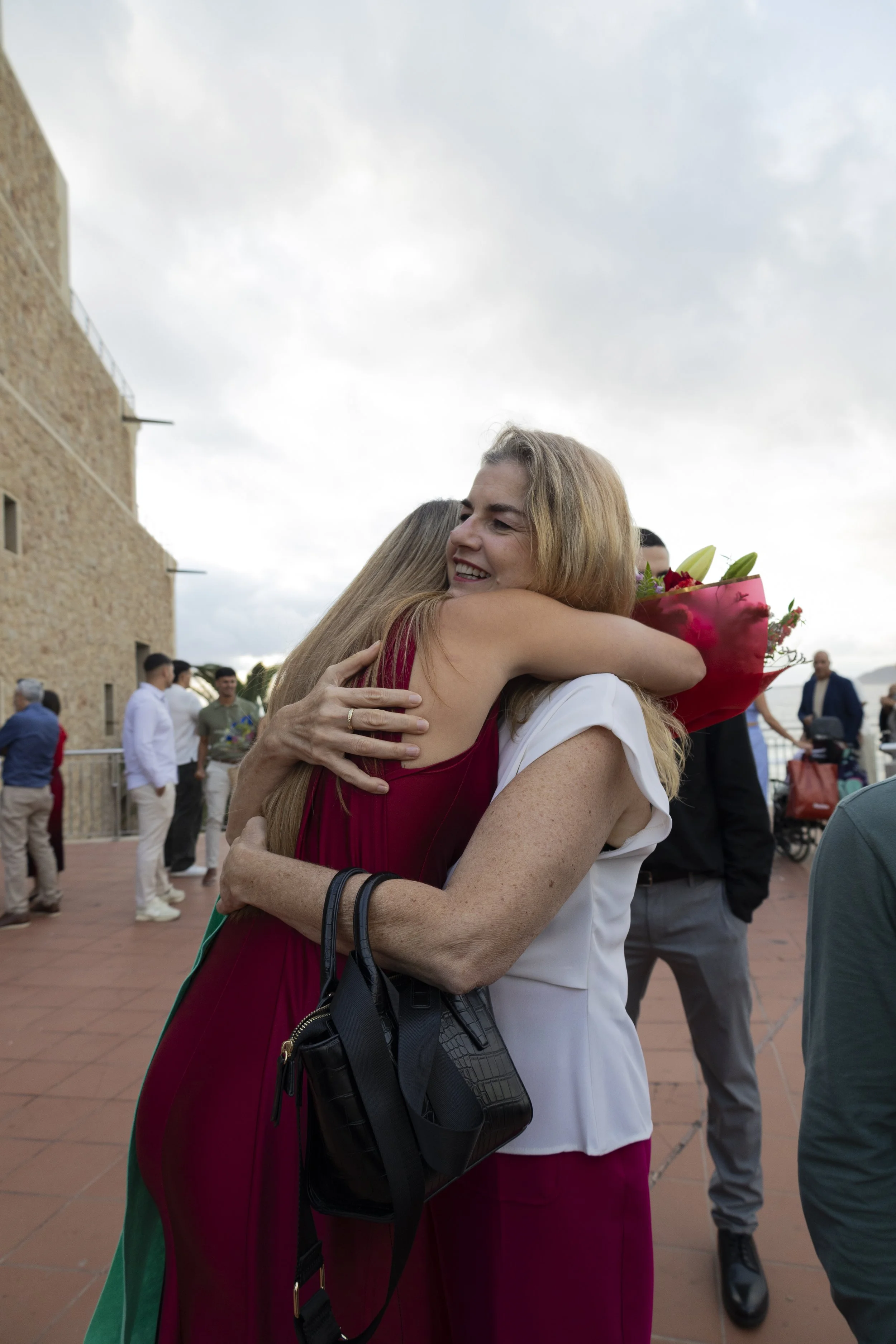 Dos mujeres se abrazan y una de ellas sostiene un ramo de flores, en un ambiente exterior con varias personas en el fondo.
