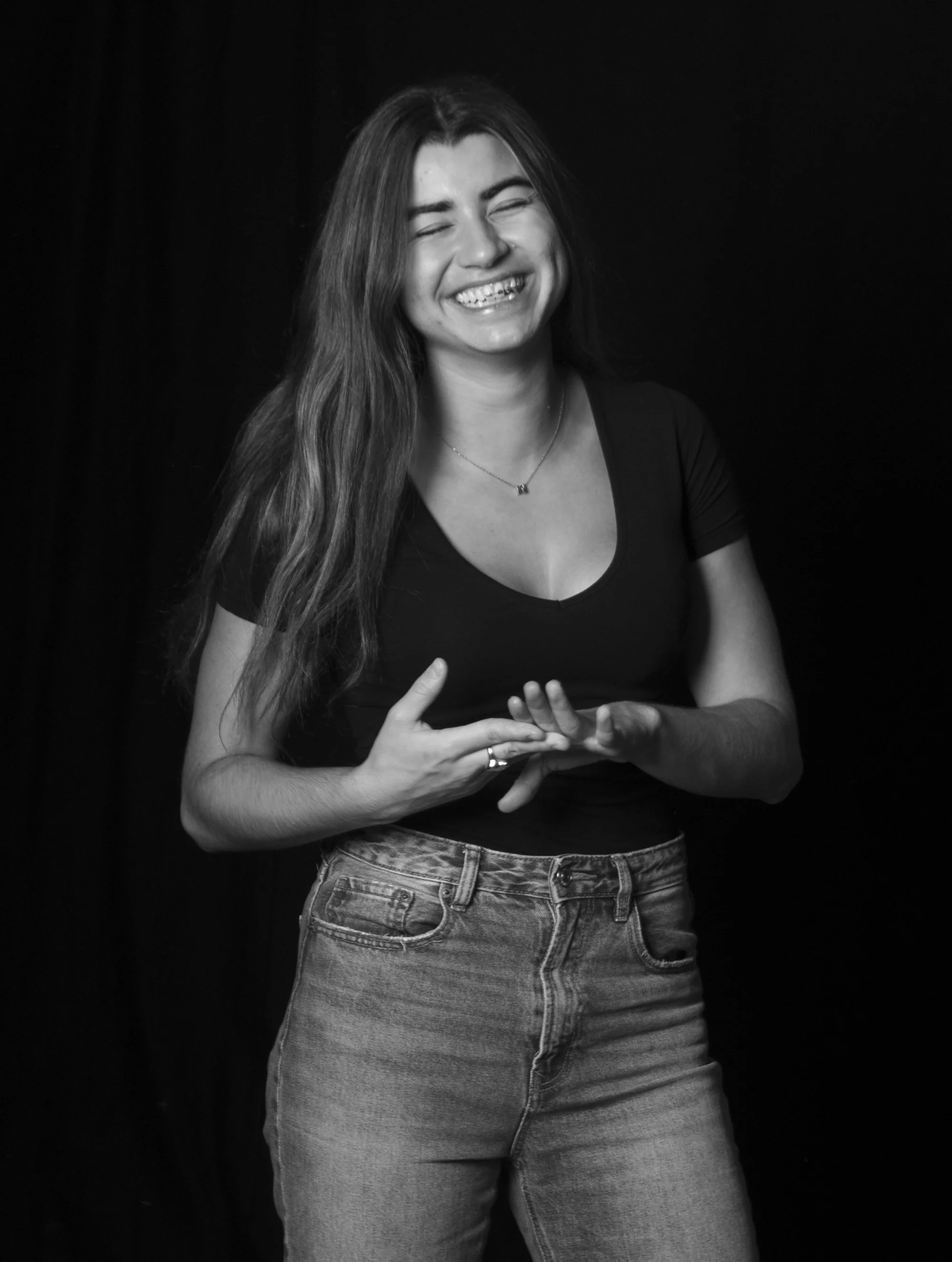 Mujer sonriendo con cabello largo, vestida con camiseta negra y jeans, en un fondo negro.