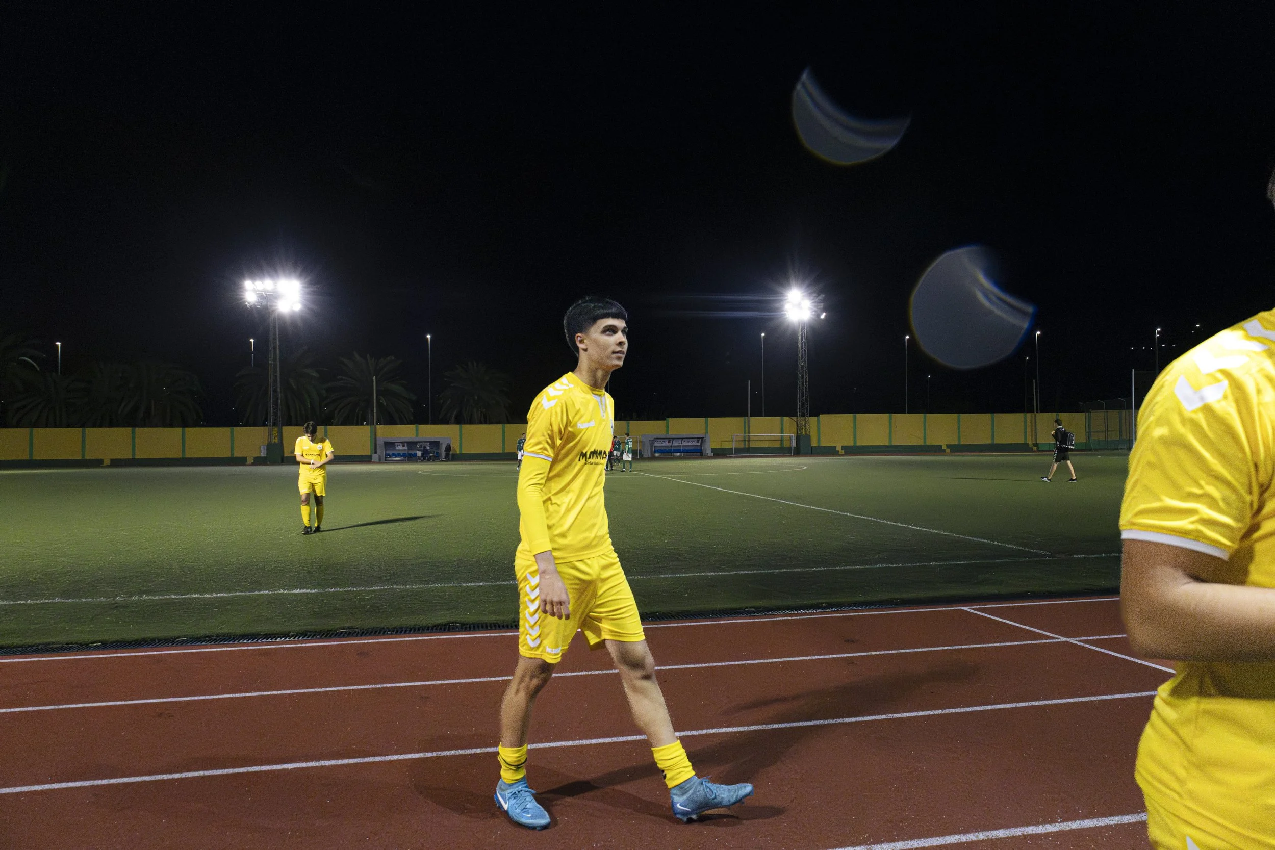 Jugadores de fútbol en uniforme amarillo en un campo iluminado por luces artificiales en la noche, en una pista de atletismo