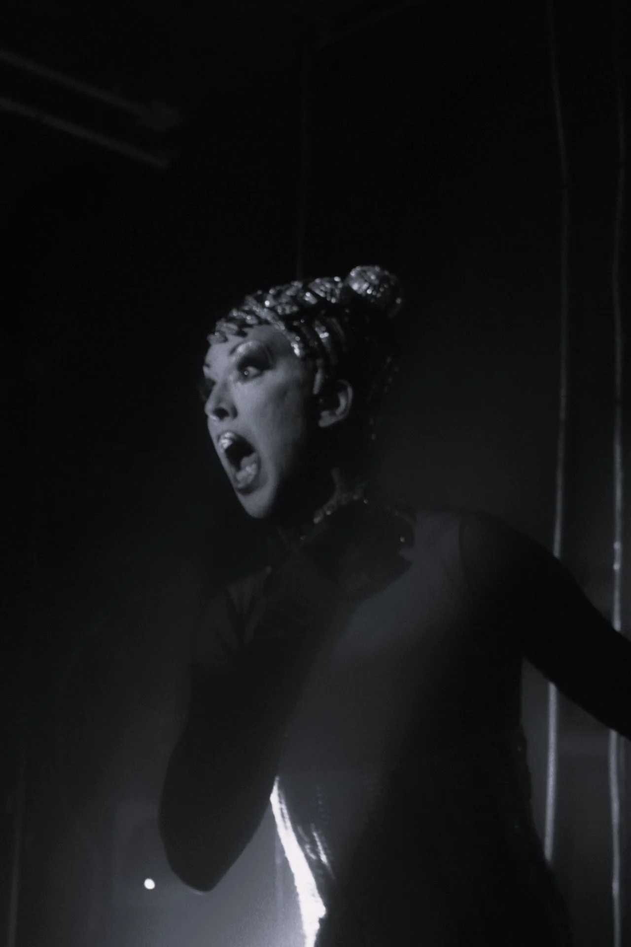 Una mujer con un peinado elaborado y maquillaje dramático, vestida con un atuendo oscuro, en una escena en blanco y negro con expresión emocional intensa.