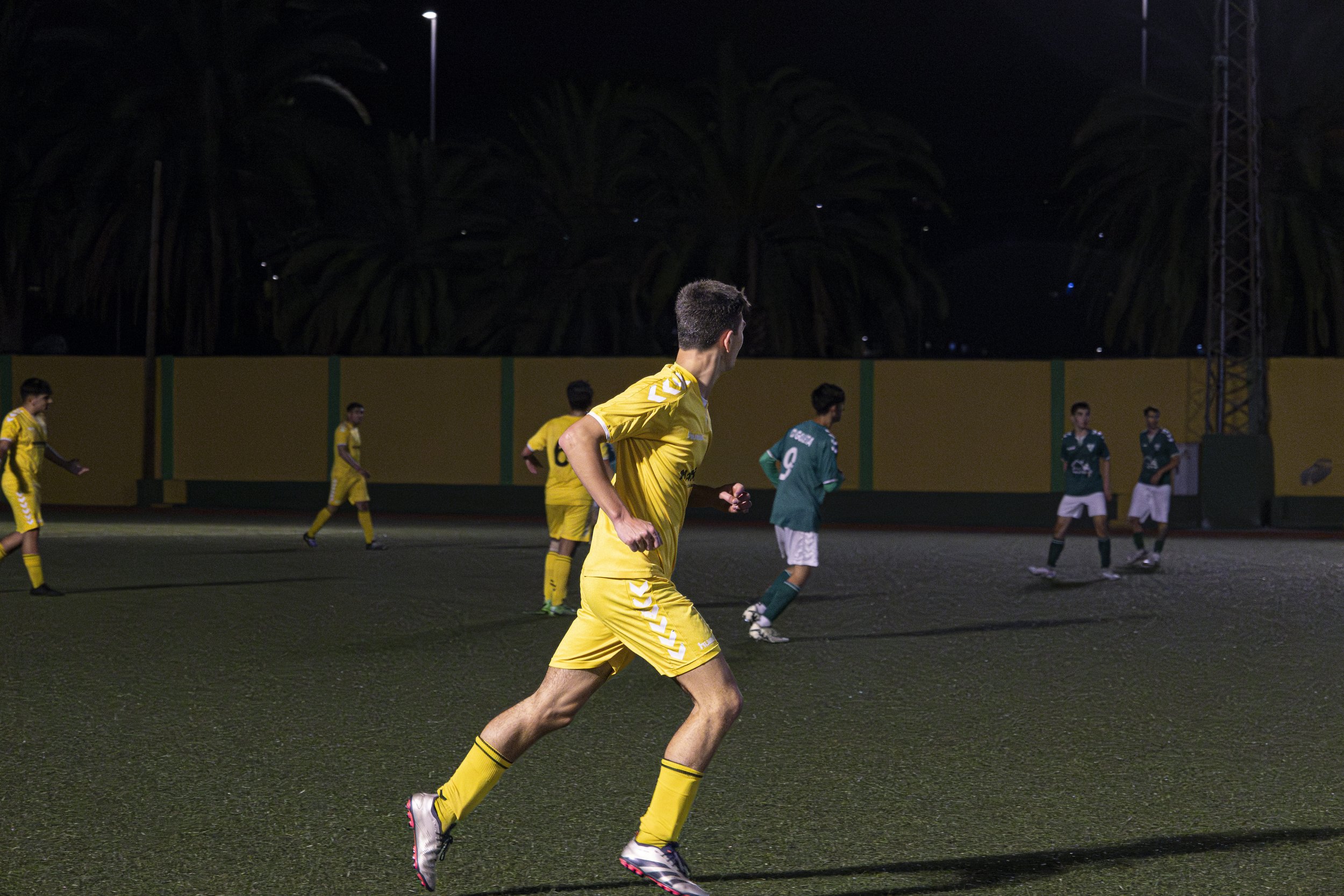 Jugadores de fútbol en un campo de noche, algunos con uniforme amarillo y otros con uniforme verde, en medio de un partido.