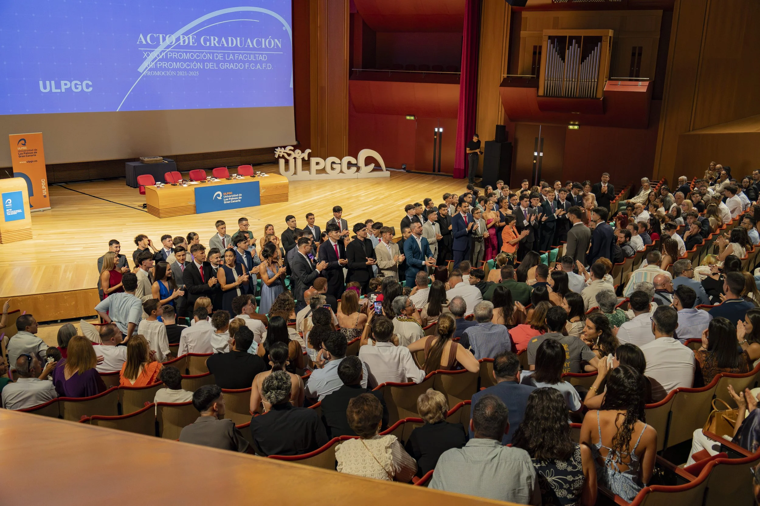 Escenario de graduación en un auditorio con muchas personas, algunos de pie y otros sentados, en la stage hay una mesa con sillas y en la pantalla grande se lee 'Acto de Graduación' y otras informaciones académicas.