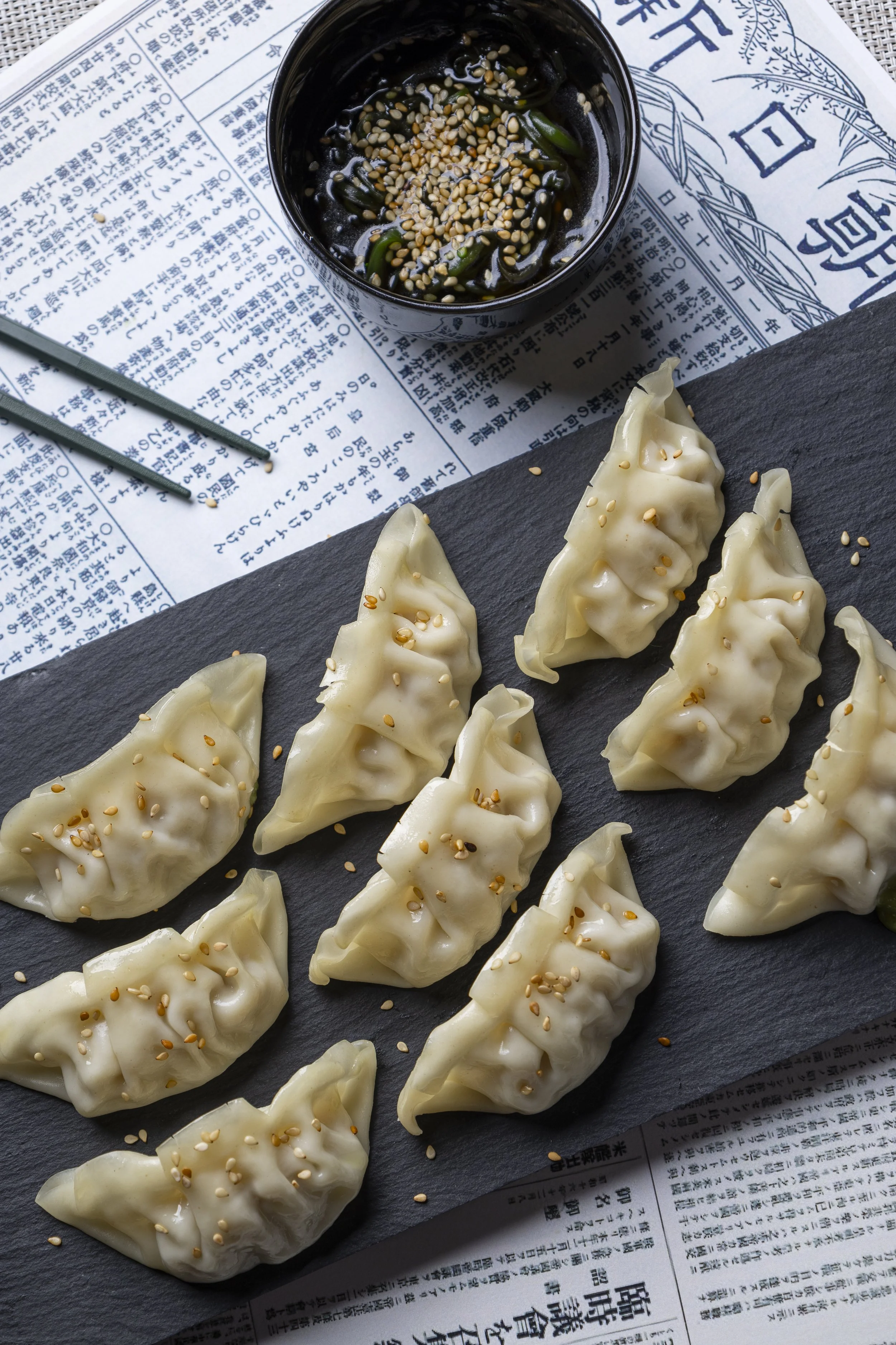Dumplings de masa blanca con semillas de sésamo doradas en un plato negro, acompañado de salsa con semillas de sésamo y cebolleta en una taza pequeña, sobre papel de periódico impreso en japonés.