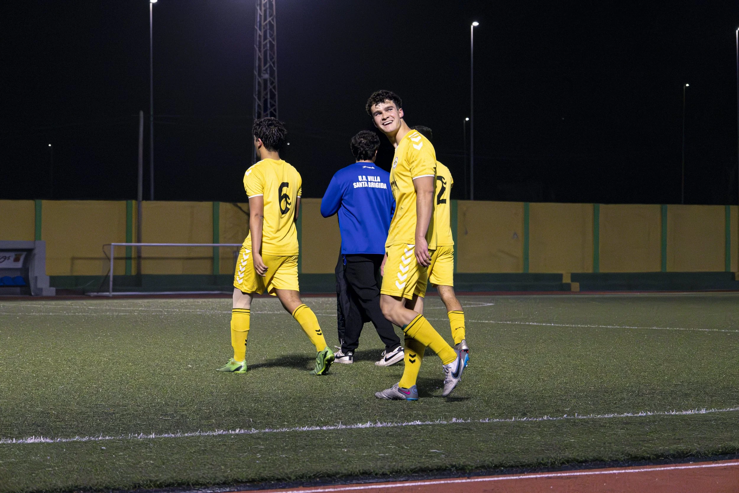 Jugadores de fútbol en el campo durante un juego nocturno, vistiendo uniformes amarillos y un asistente con chaqueta azul.
