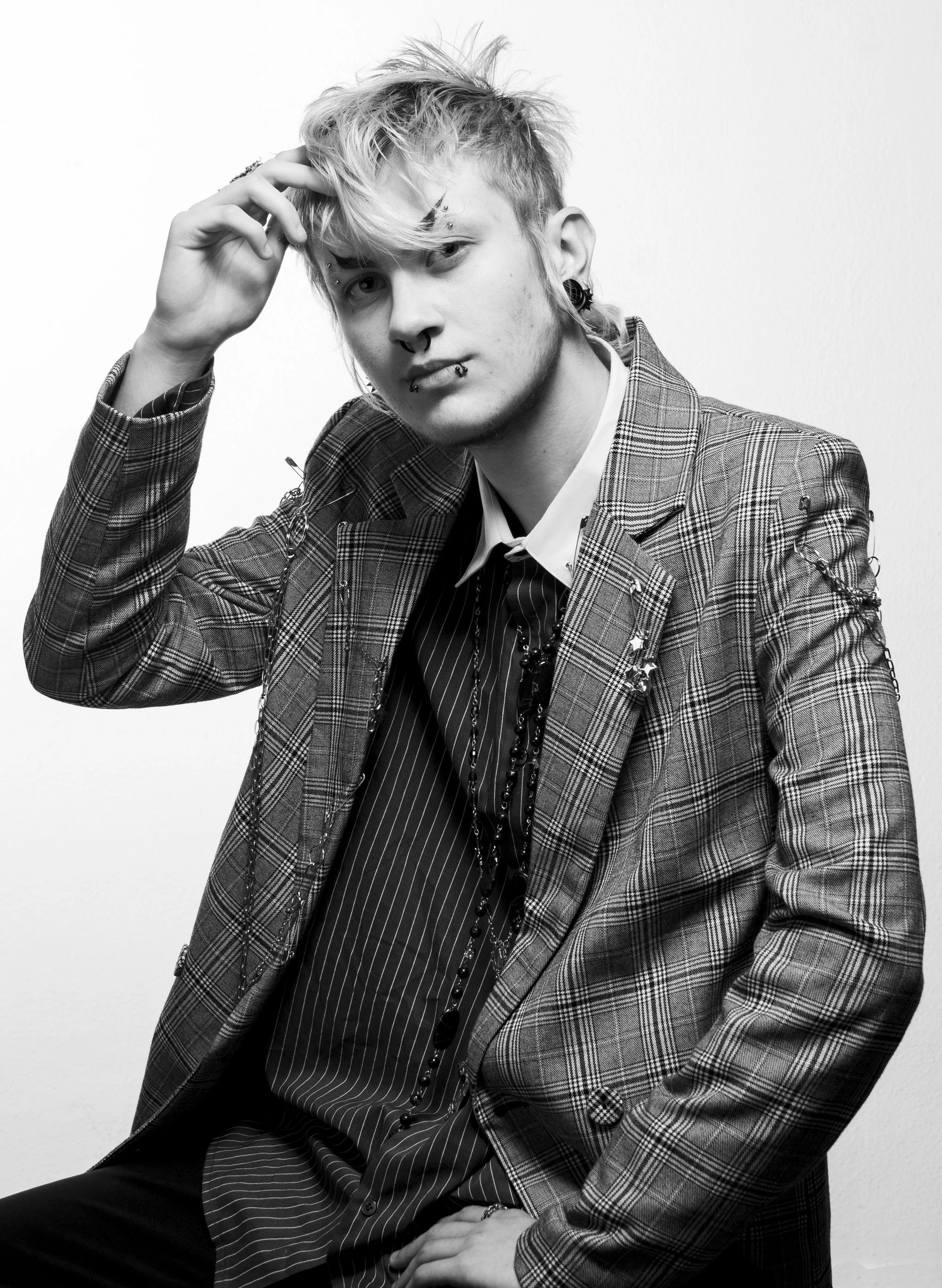 Joven con cabello despeinado, piercings y tatuajes, vistiendo traje a cuadros y ropa elegante, posando en un fondo blanco.