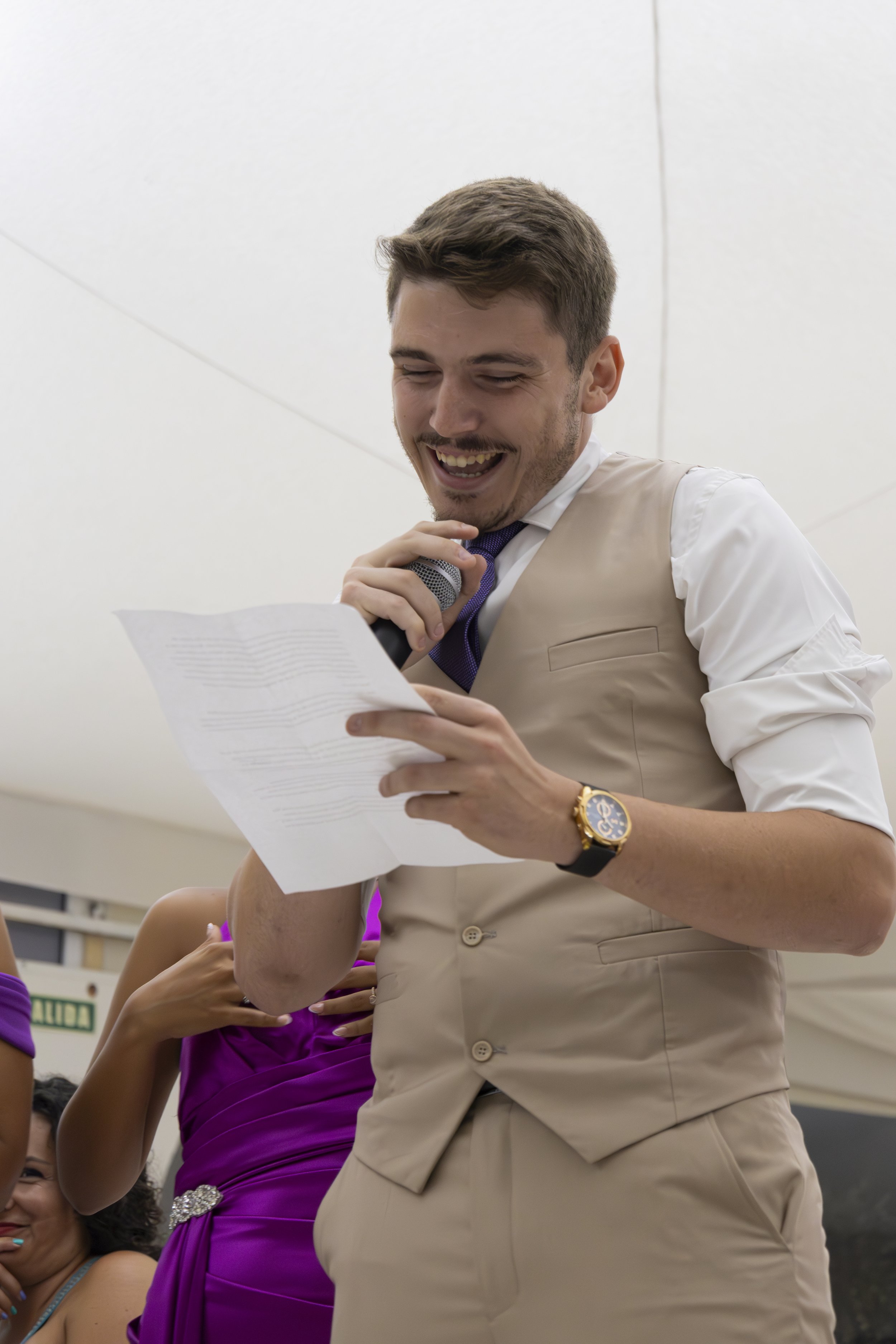 Un hombre sonriendo lee un papel mientras sostiene un micrófono, vestido con camisa blanca, chaleco beige y reloj dorado.