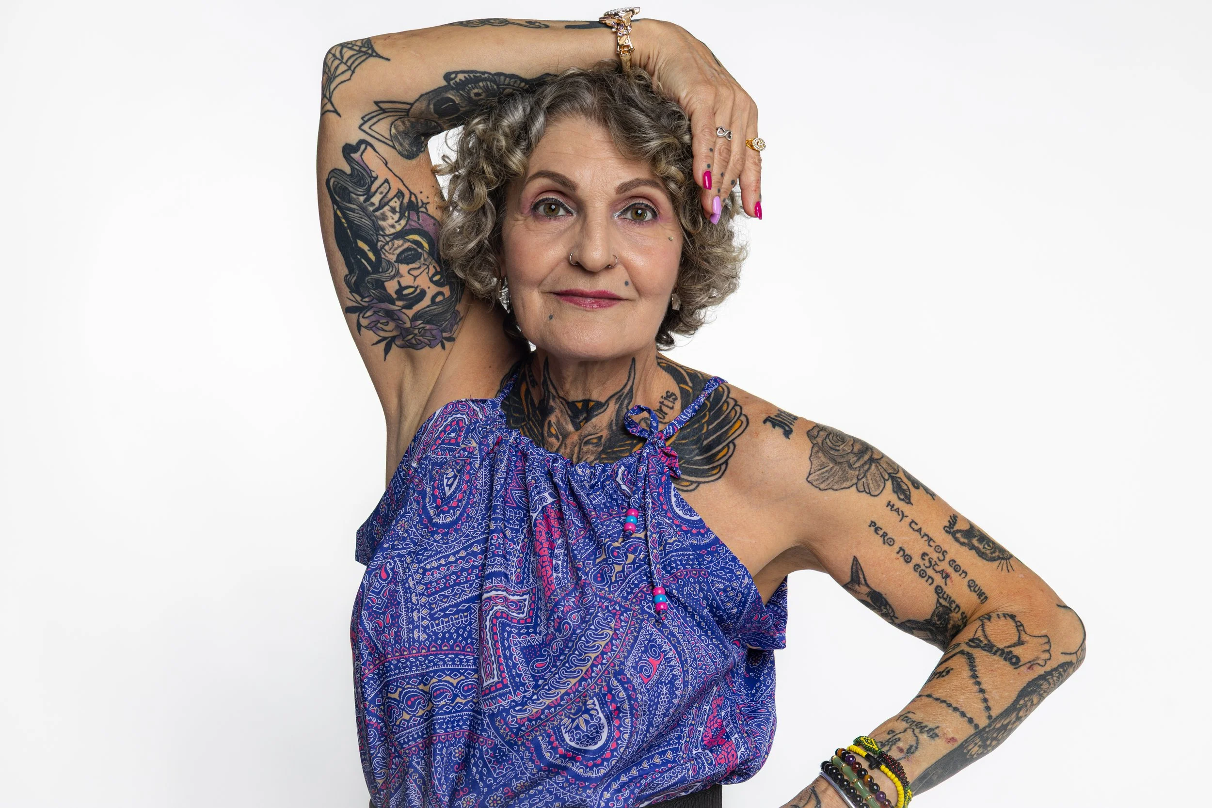 Una mujer mayor con cabello rizado y canoso, con múltiples tatuajes en brazos, cuello y rostro, lleva un vestido azul con detalles en rosa y negro, y adorna con pulseras de colores, posando con una mano en la cabeza contra un fondo blanco.