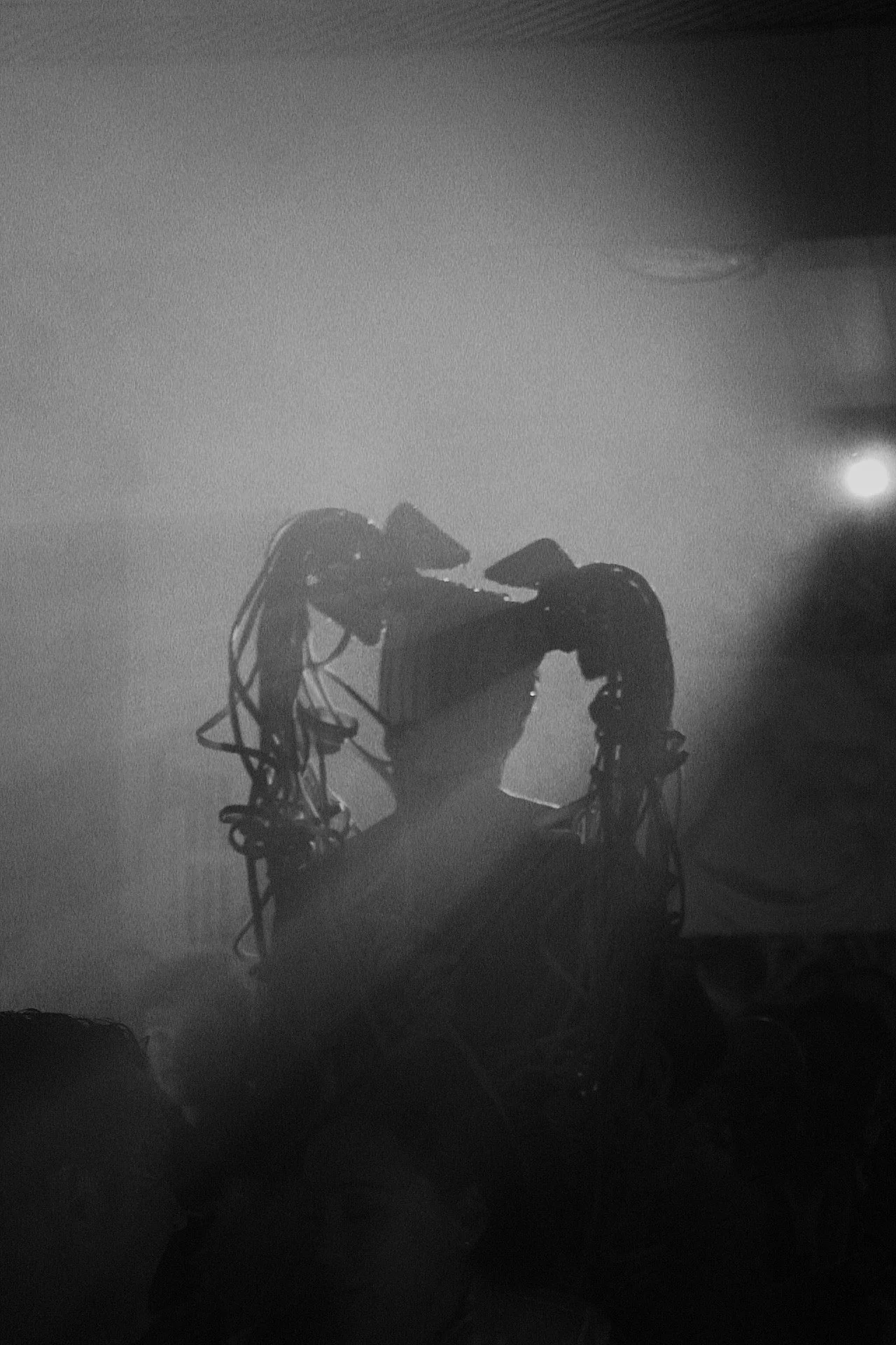 Fotografía en blanco y negro de un robot o máquina con cables y estructuras metálicas que sostienen una caja o caja fuerte, con una luz brillante en el fondo