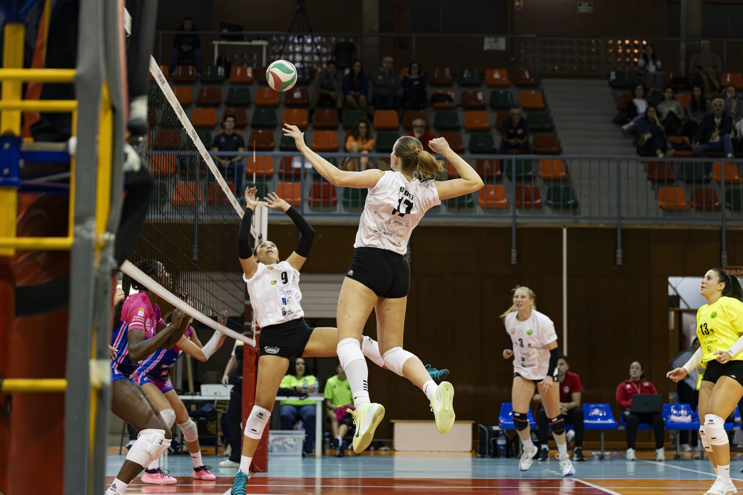 Juego de voleibol en acción en una cancha cubierta, con jugadoras en movimiento y espectadores observando desde las gradas.