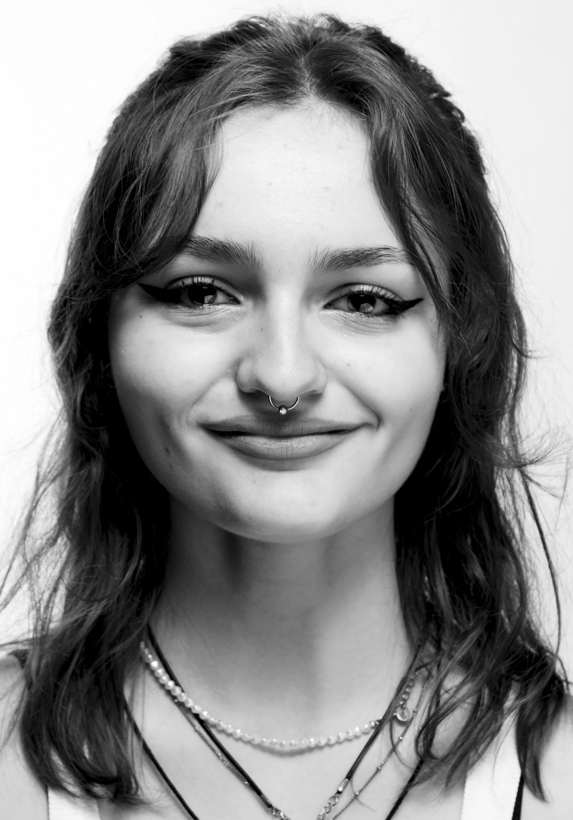 Retrato en blanco y negro de una joven con cabello rizado, sonrisa suave, piercings en la nariz, y varias cadenas en el cuello.