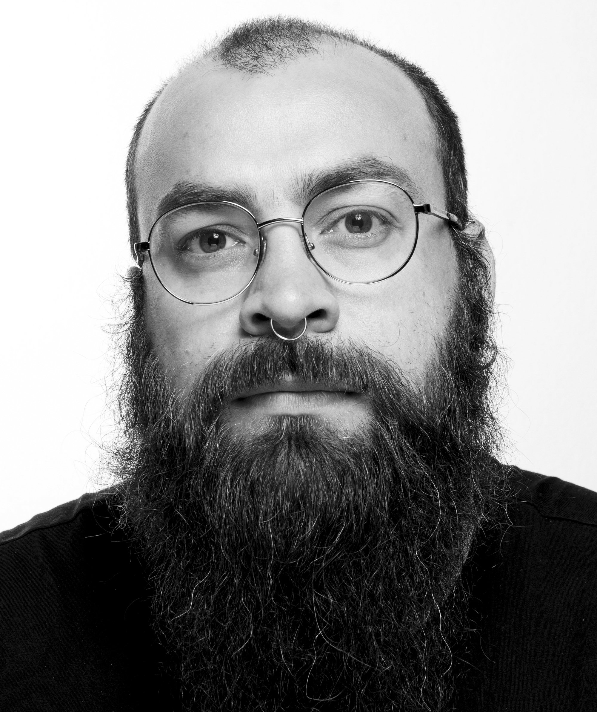 Retrato en blanco y negro de un hombre con barba, gafas redondas y septum piercing, mirando hacia la cámara.