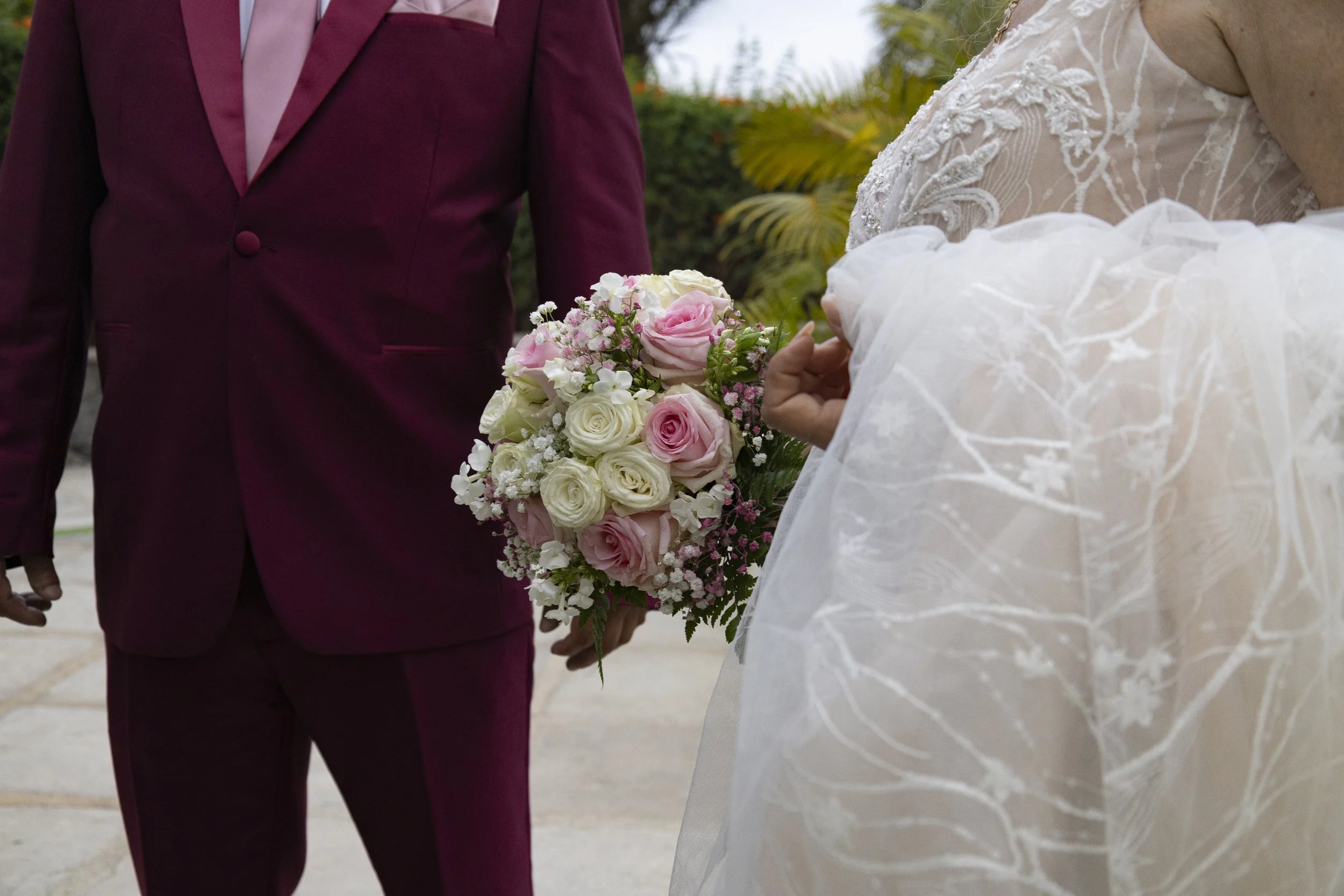 Pareja en Ibiza en una boda, la mujer con vestido y el hombre con traje burdeos, ella sostiene un ramo de flores rosadas y blancas.