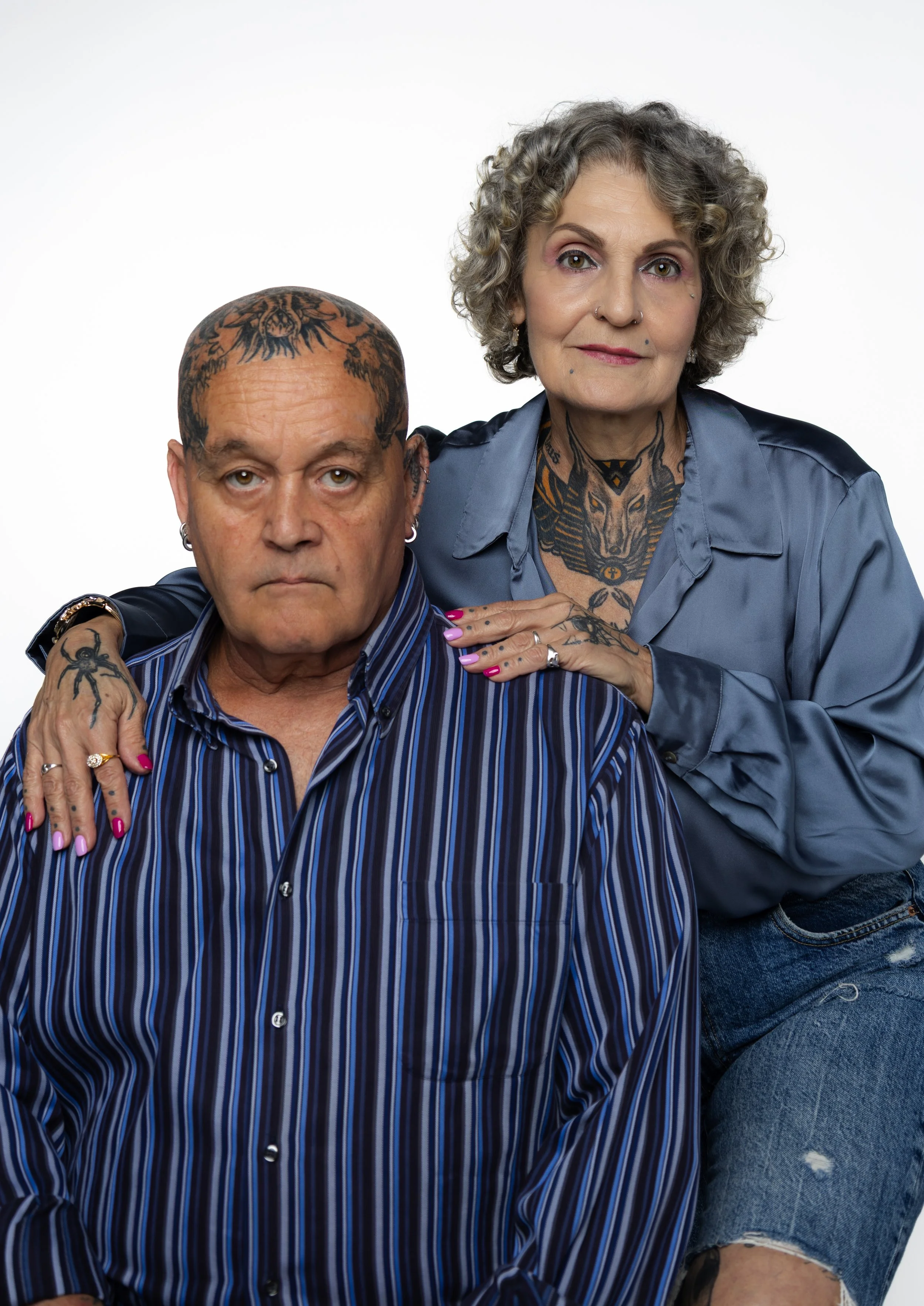 Un hombre y una mujer con tatuajes, ambos con camisetas a rayas, posando juntos contra fondo blanco.
