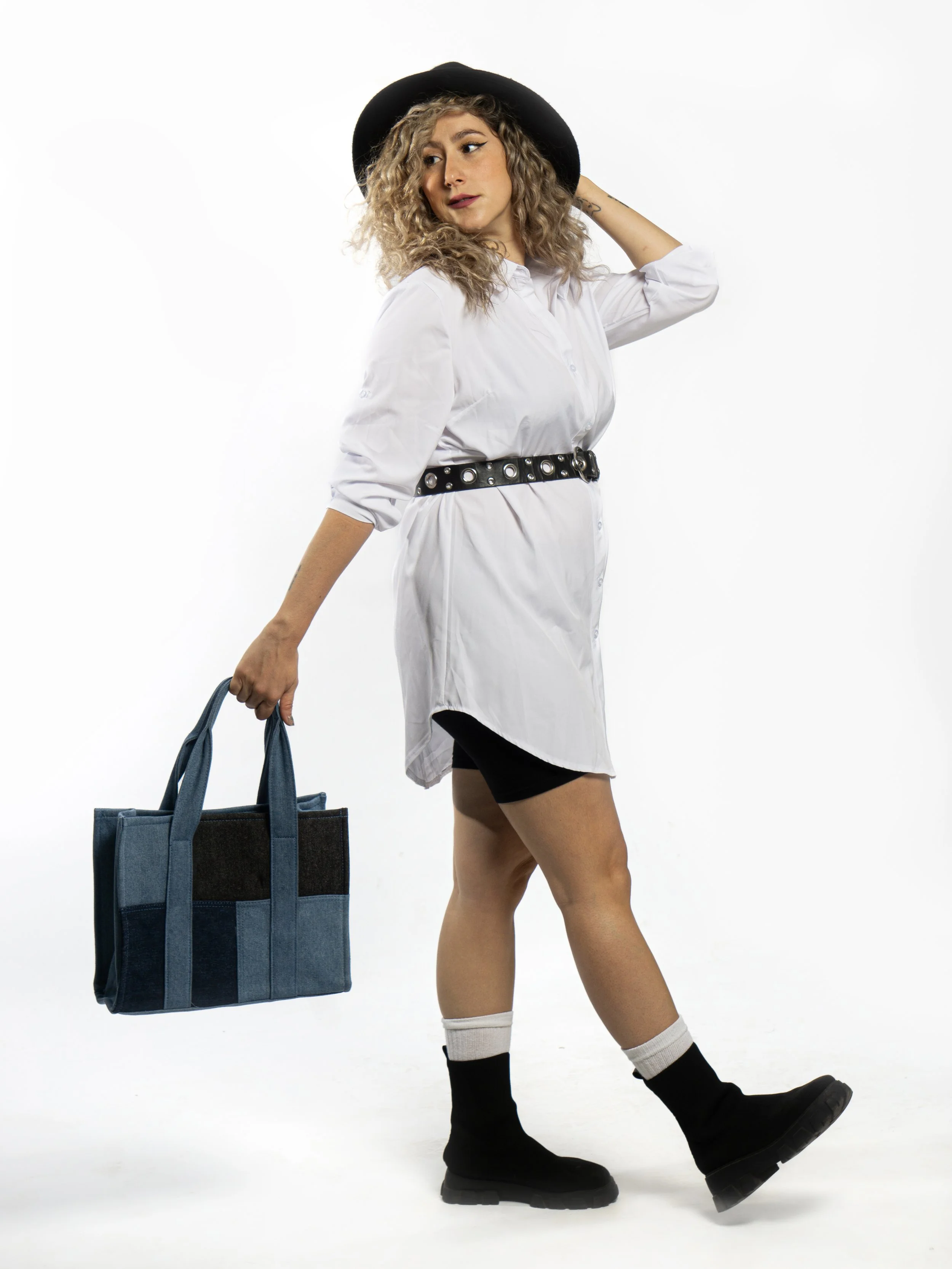 Mujer con cabello rizado rubio, sombrero negro, blusa blanca, cinturilla negra con ojales, pantalones cortos negros, medias blancas y botas negras, sosteniendo una bolsa grande de mezclilla, en un fondo blanco.