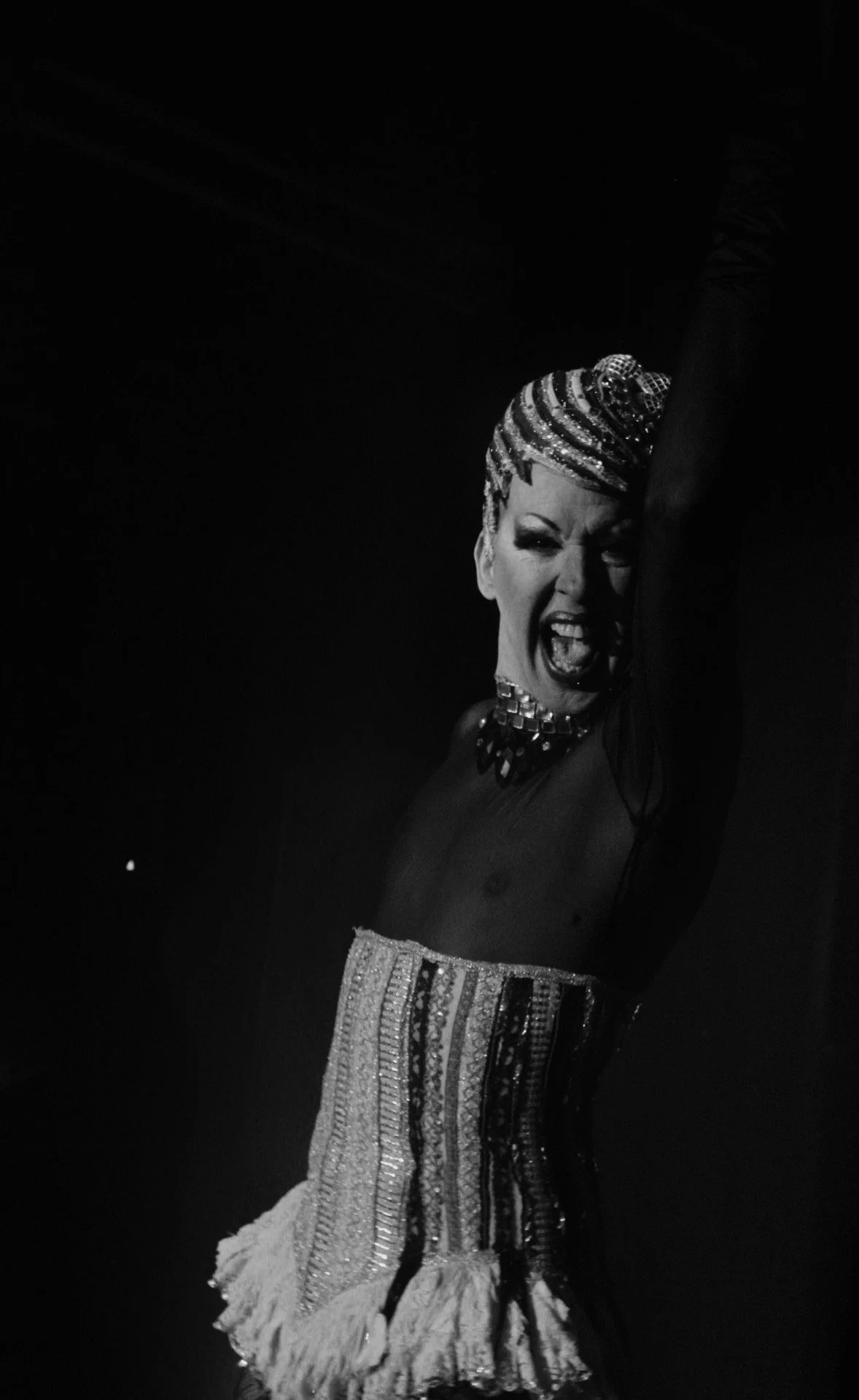 Una mujer en escenario con expresión de emoción, llevando accesorios y ropa con detalles brillantes, en un efecto en blanco y negro.