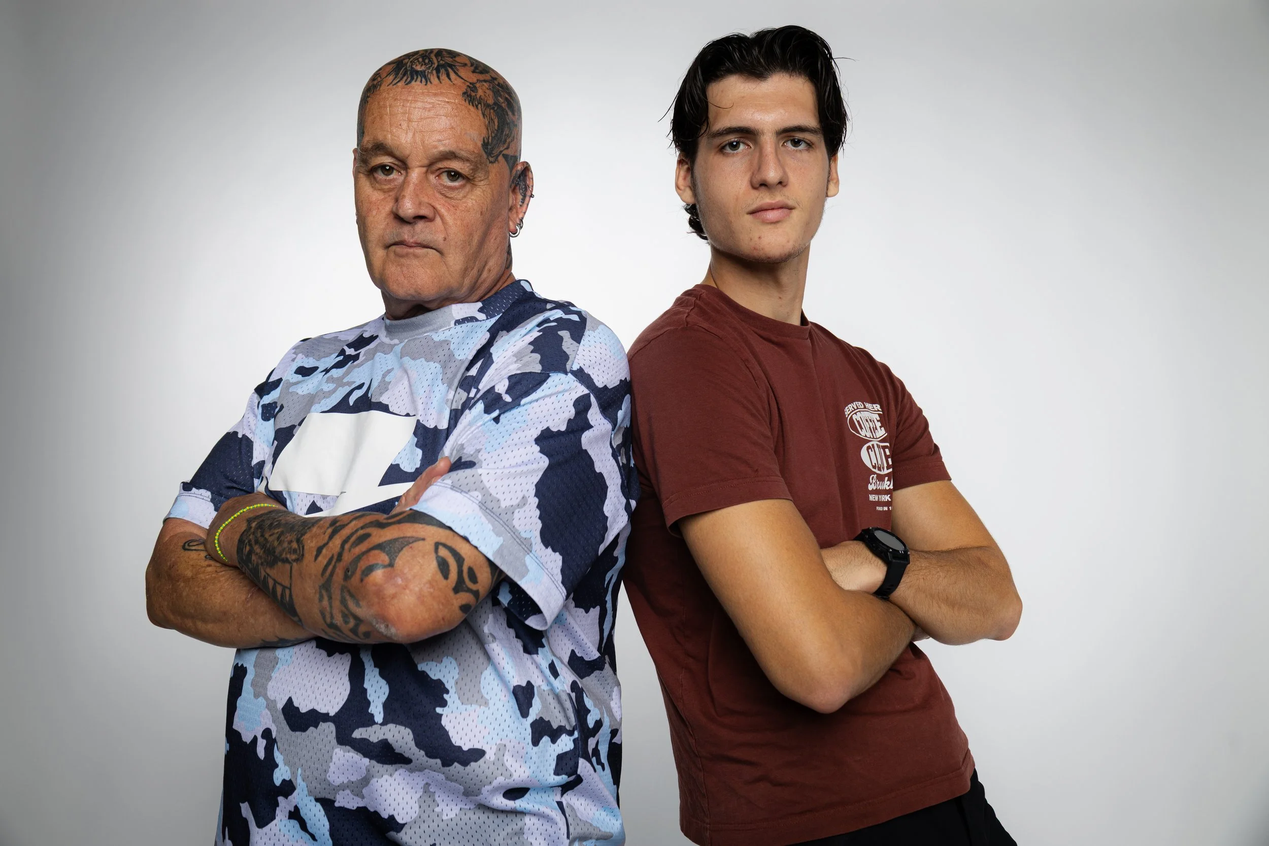 Dos hombres con los brazos cruzados y expresión seria, uno con tatuajes y camiseta de camuflaje, otro con camiseta roja y reloj, de pie en fondo gris.