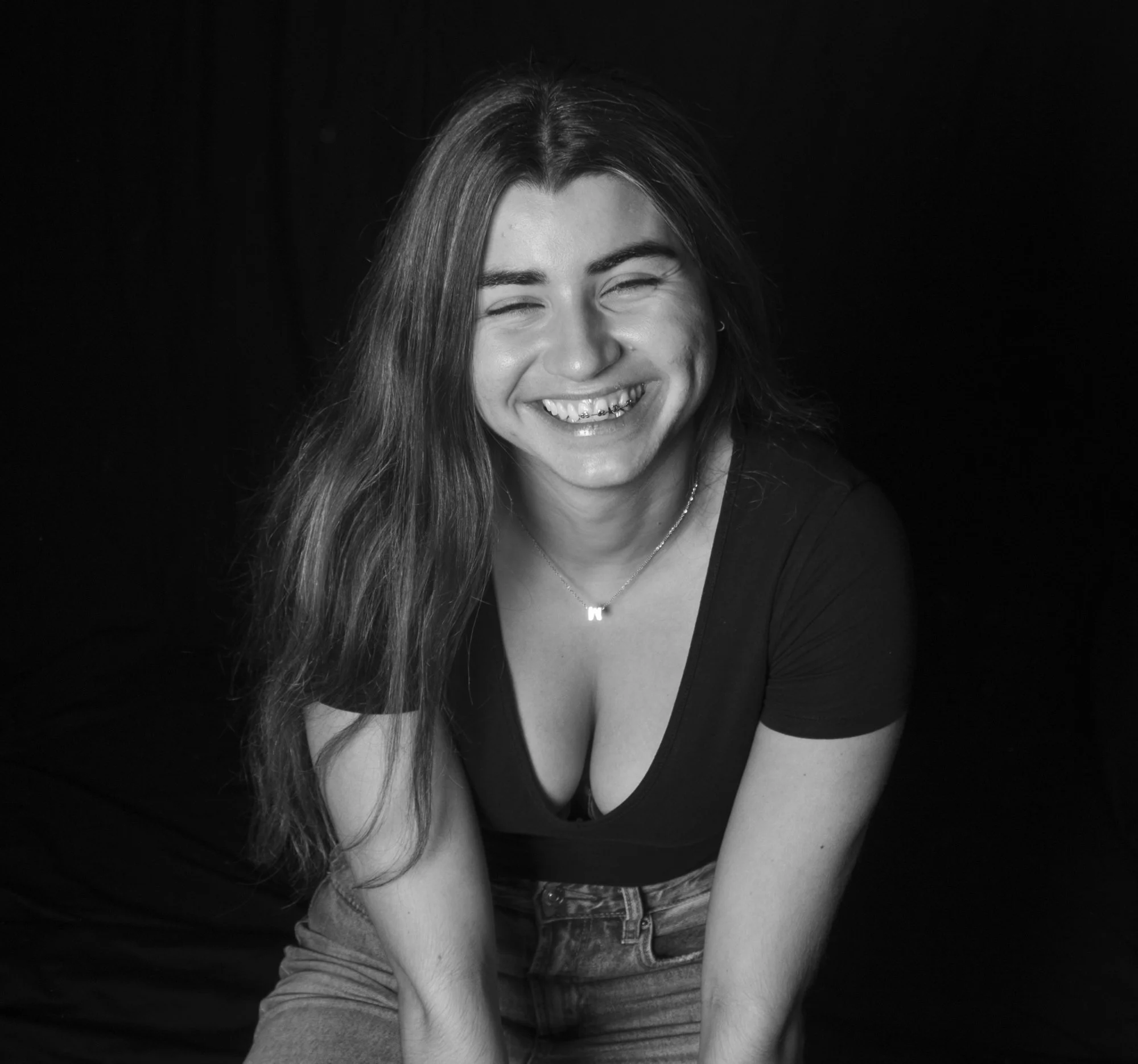 Retrato en blanco y negro de una mujer sonriendo con cabello largo y oscuro, usando camiseta negra y collar, contra fondo negro.