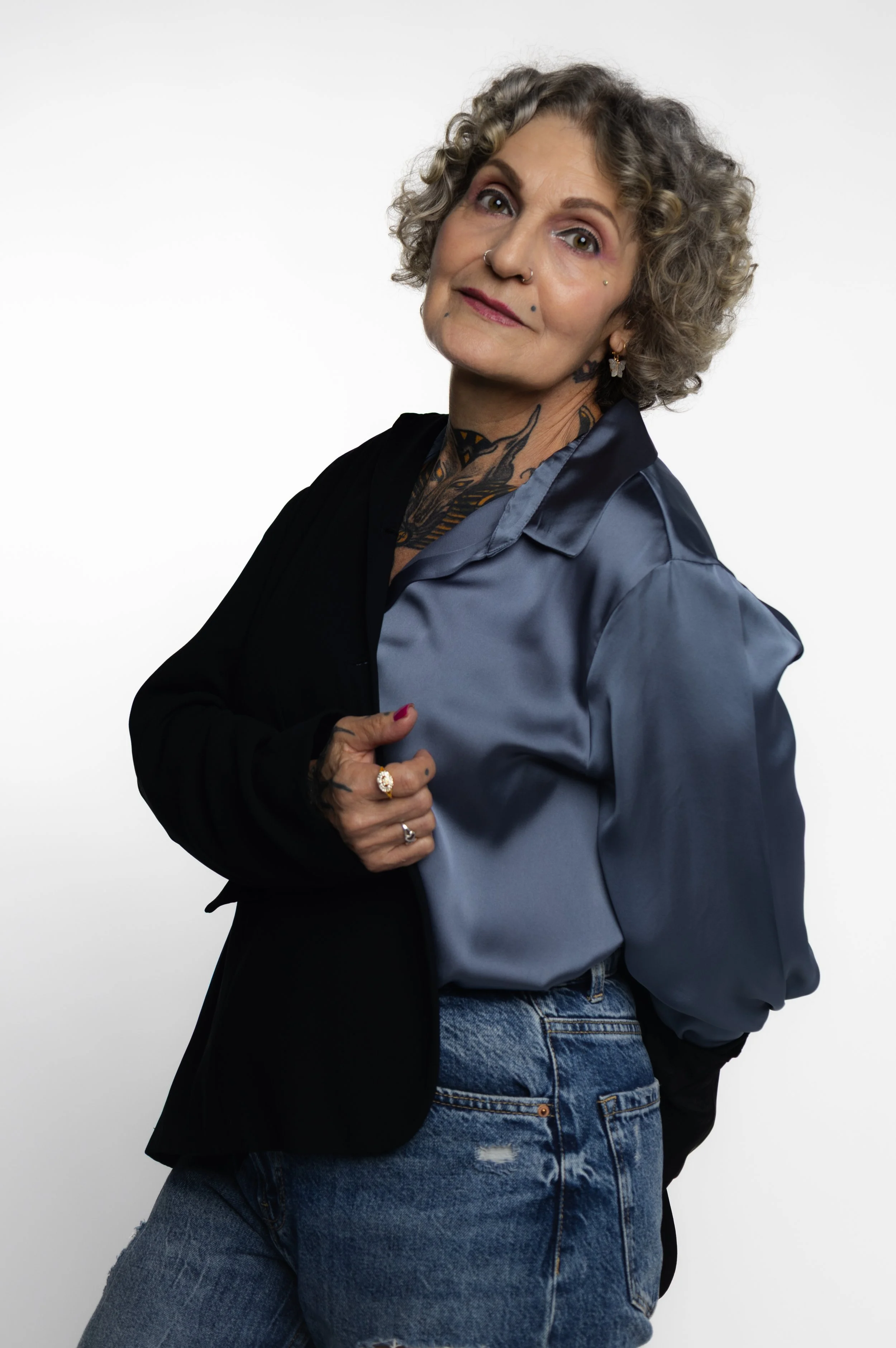 Mujer con cabello rizado, tatuajes, vestida con camisa de satén azul y pantalones vaqueros, posando en estudio con fondo blanco.