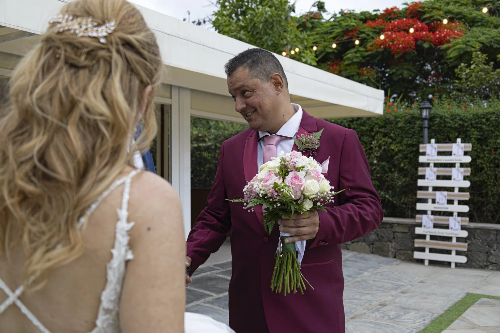 Un hombre con traje burdeos entrega un ramo de flores a una mujer en un entorno al aire libre, con árboles y decoraciones florales.