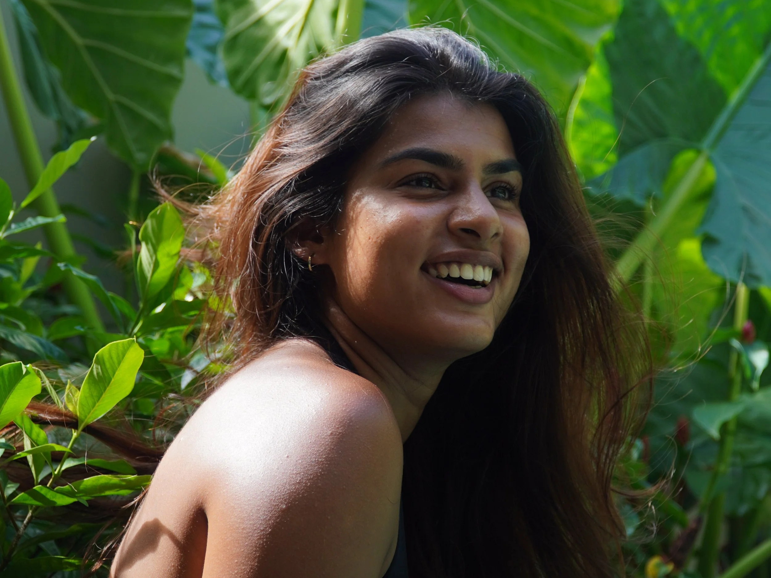 A woman with long, dark hair smiling amidst lush green tropical plants.