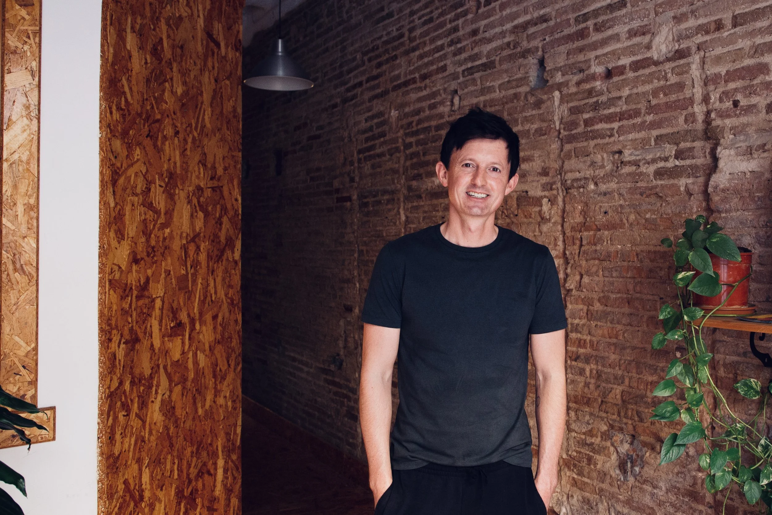 Smiling man with black hair wearing a black t-shirt standing against a brick wall interior with green plants and wooden accents.