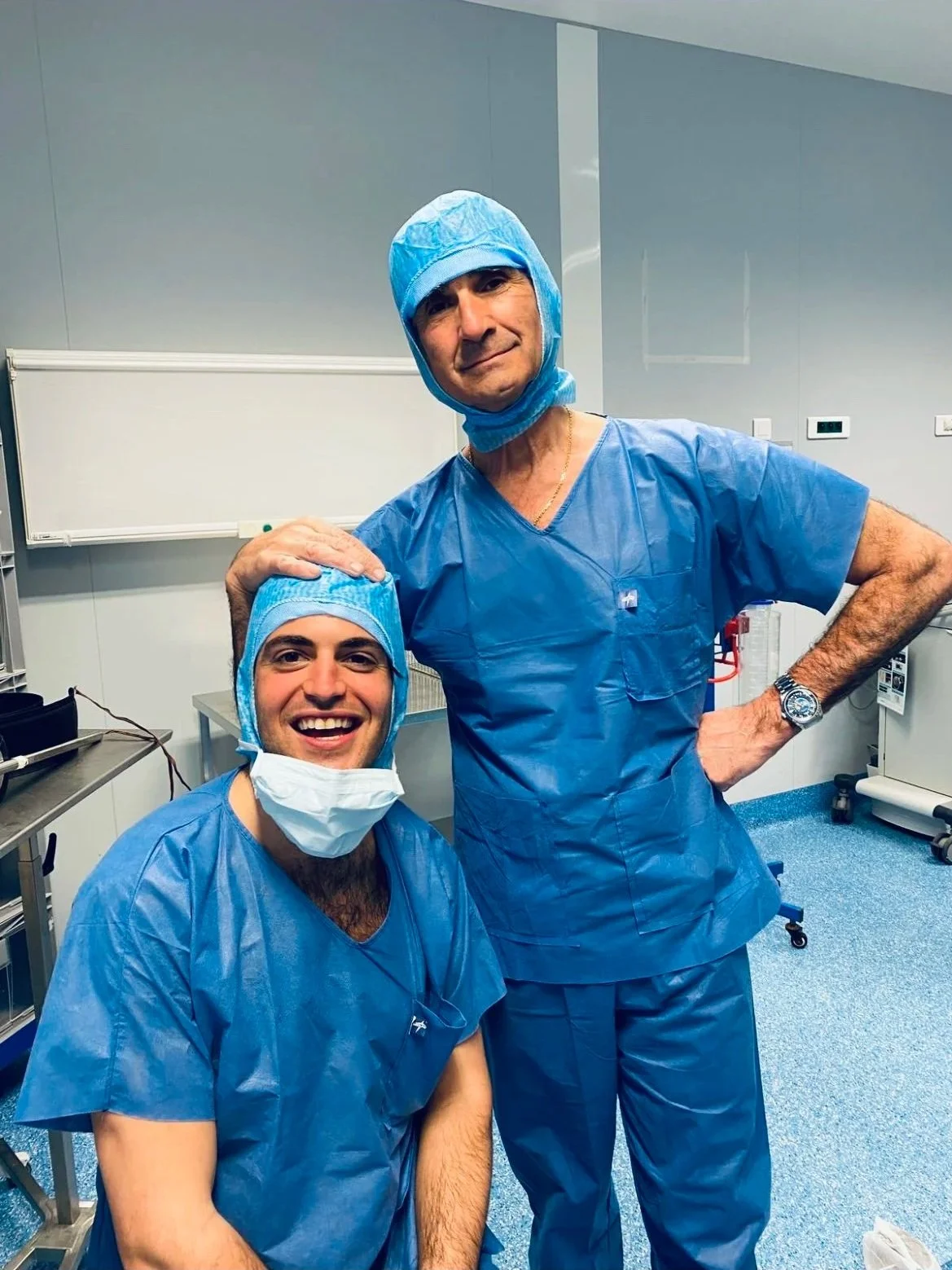 Two male medical professionals in scrubs and surgical caps posing in an operating room, one sitting and smiling, the other standing with hand on his hip.