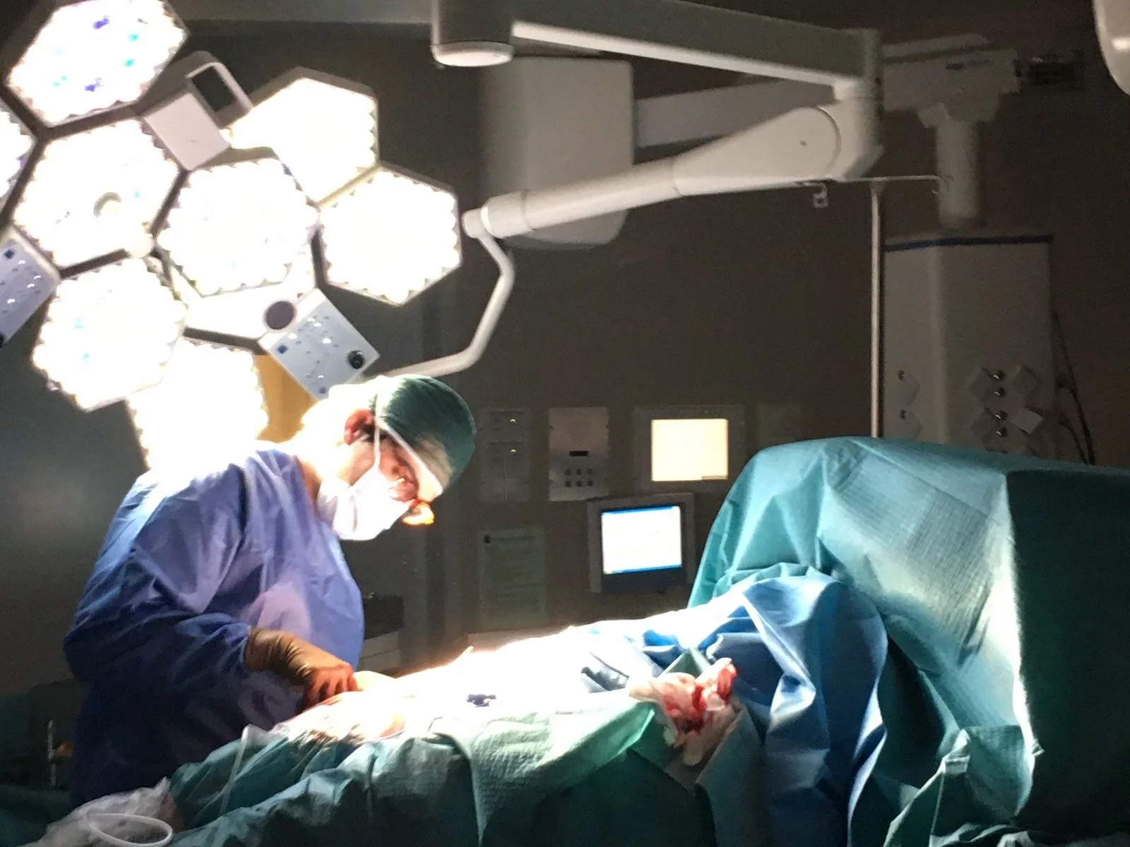 A surgeon wearing scrubs, mask, and protective eyewear operating in an operating room. Surgical lights are overhead. The patient is covered with a surgical drape.